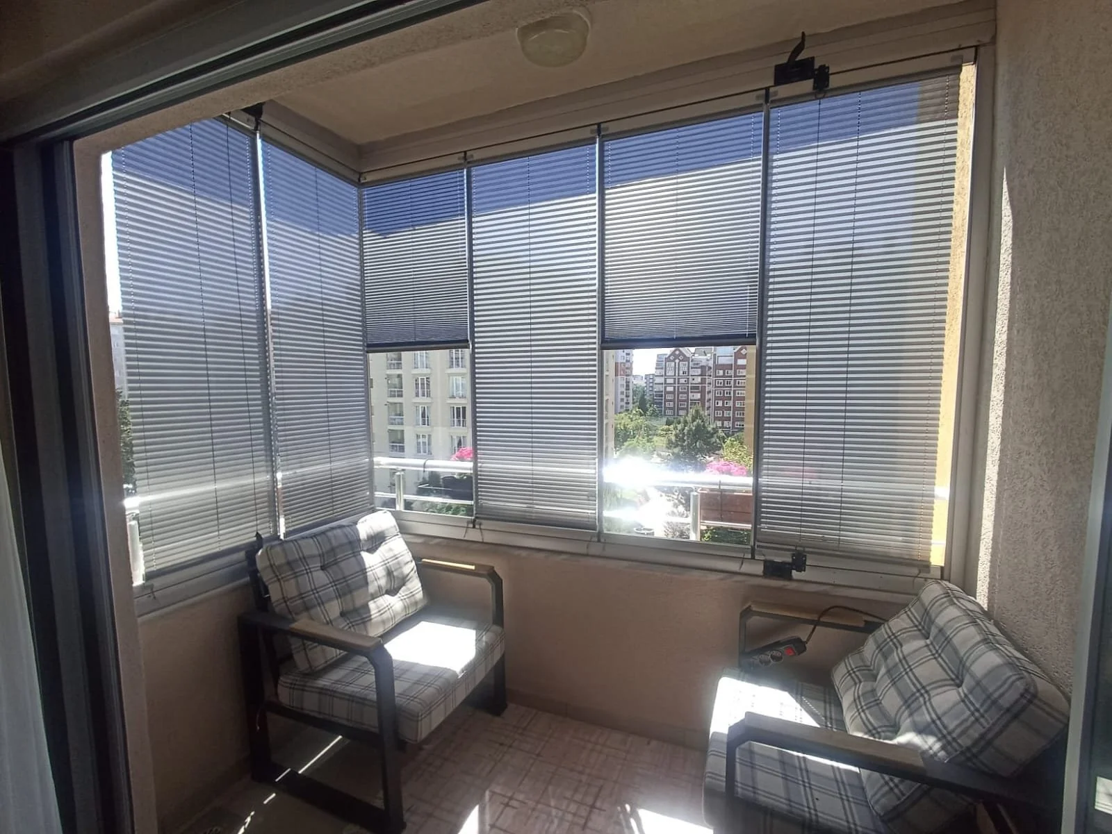 A balcony with windows covered by horizontal blinds, two plaid armchairs, and a view of nearby apartment buildings and greenery outside.