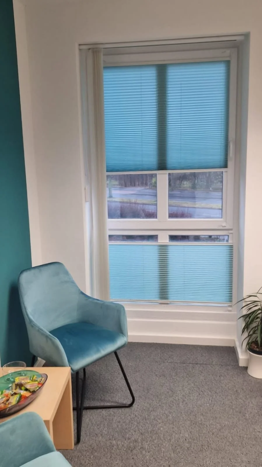 A cozy office corner with a light blue velvet armchair, a small wooden side table with snacks, a window with partially closed blinds, and a potted plant on the right.