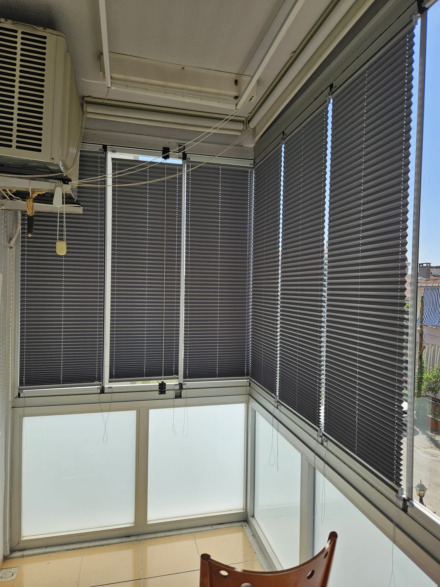 A small enclosed balcony with large windows covered by closed black horizontal blinds, a beige tiled floor, an air conditioning unit, and a wooden chair in the corner.