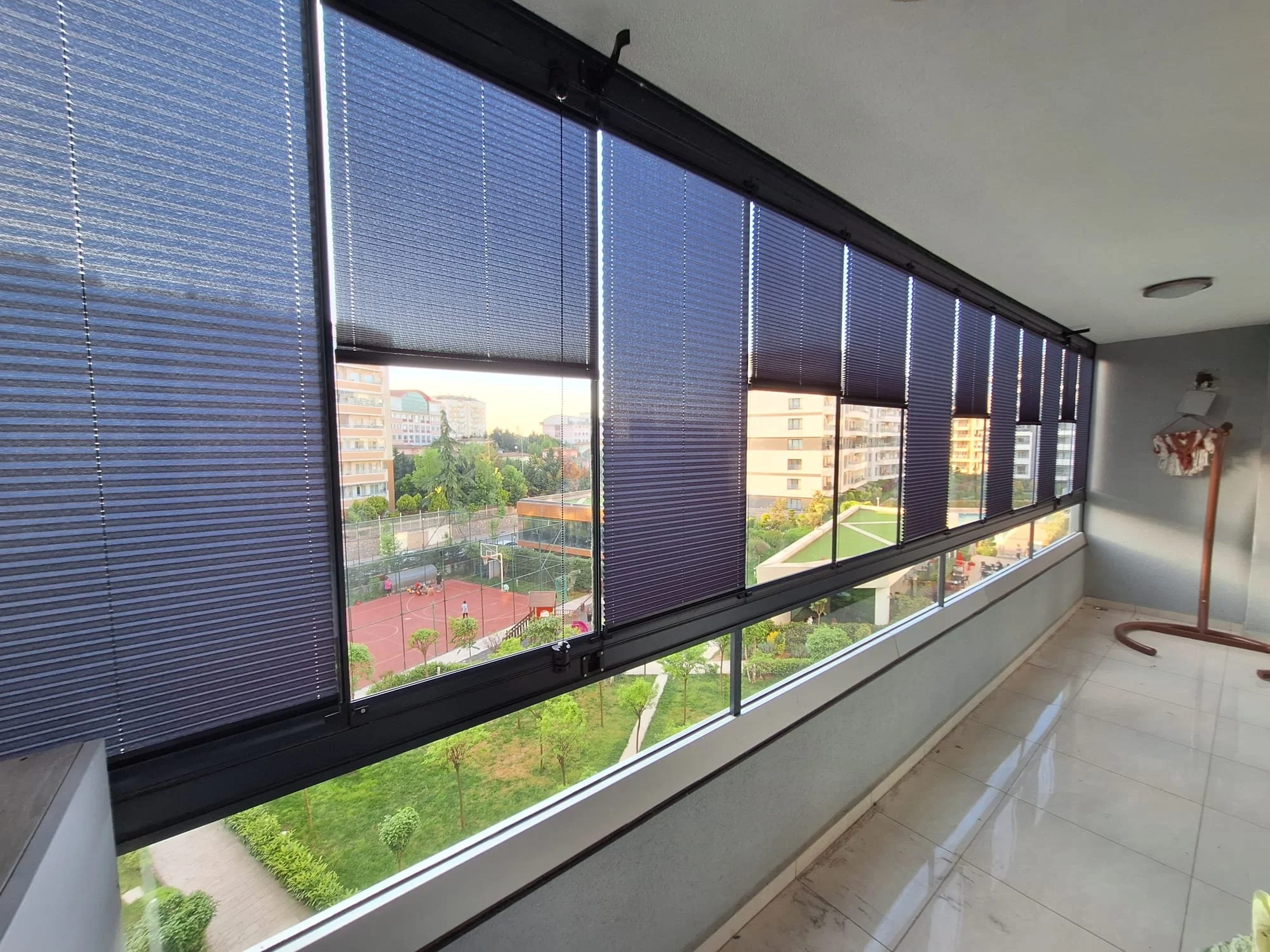 A view through large windows with blue horizontal blinds, showing a green outdoor area with trees, a sports court with basketball hoops, and adjacent residential buildings at sunset.