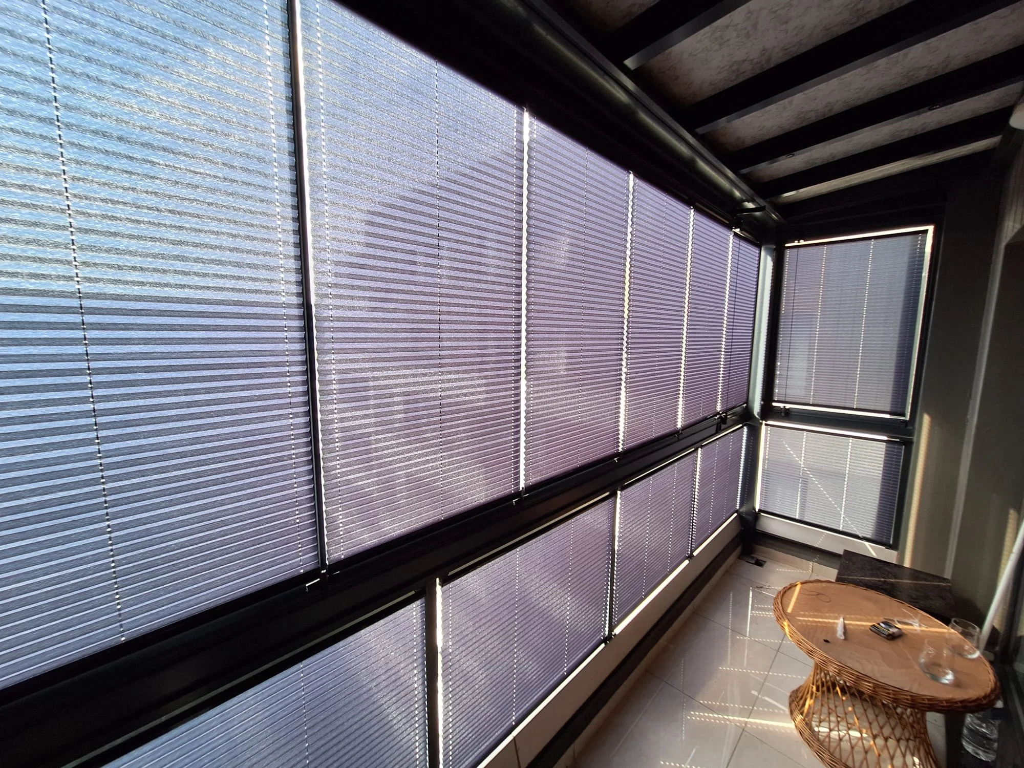 Sunlit room with large windows covered with horizontal blinds, a small wicker table with a water bottle and keys, and a tiled floor.