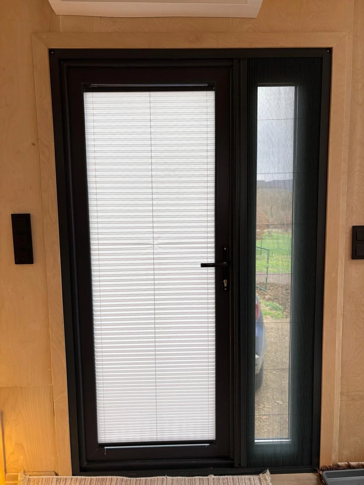 A black framed front door with a large glass panel covered by white horizontal blinds and a smaller side panel with a glass window. The door handle and lock are visible.
