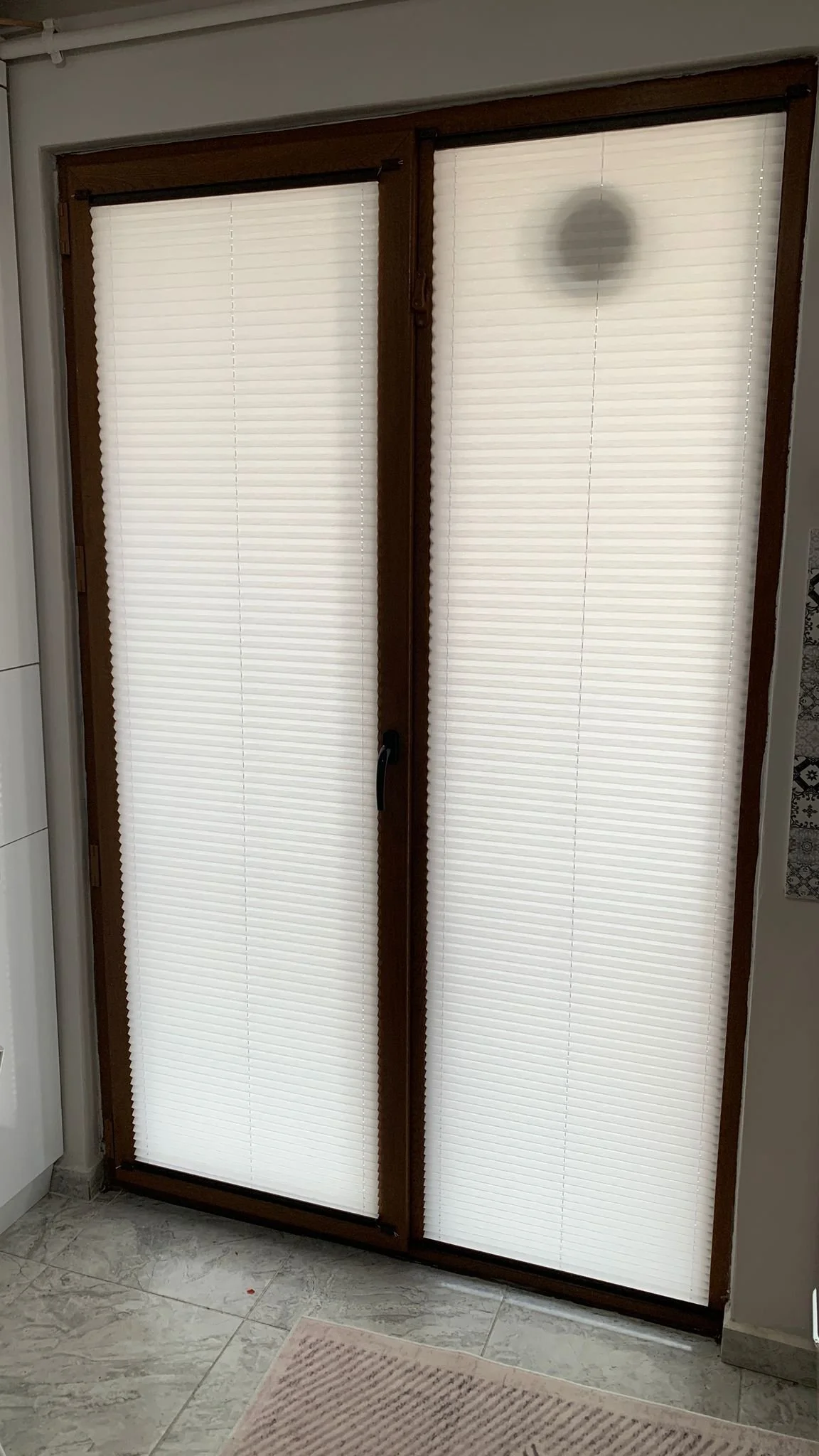 Double sliding glass doors covered with white pleated blinds, framed with dark wood, located in a room with light-colored tiled floor and part of a beige rug in front.