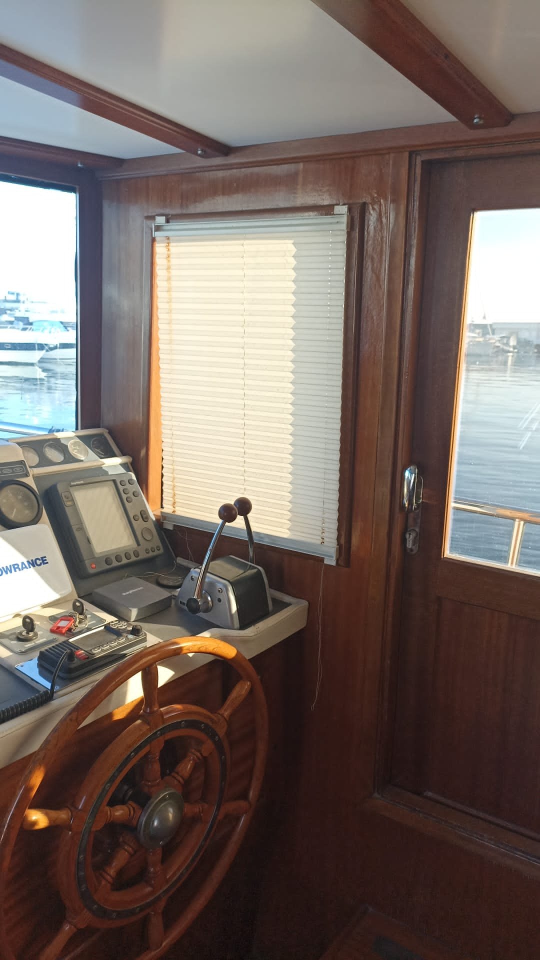 Navigation and communication equipment on a boat helm station with wooden interior, window blinds partially closed, and view of boats docked outside.