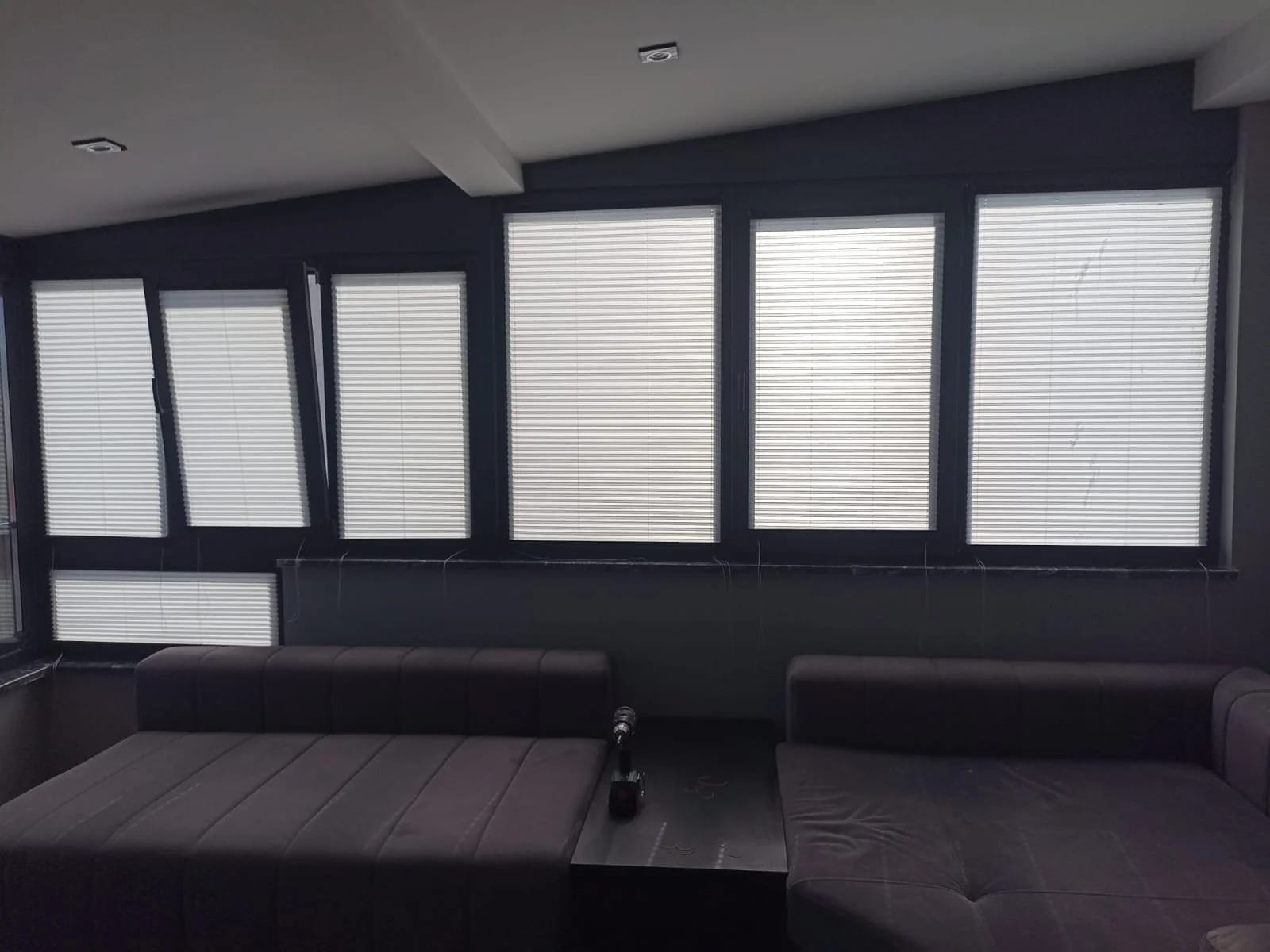 A room with dark walls and large windows with blinds, and a gray couch with a small table in front of it.