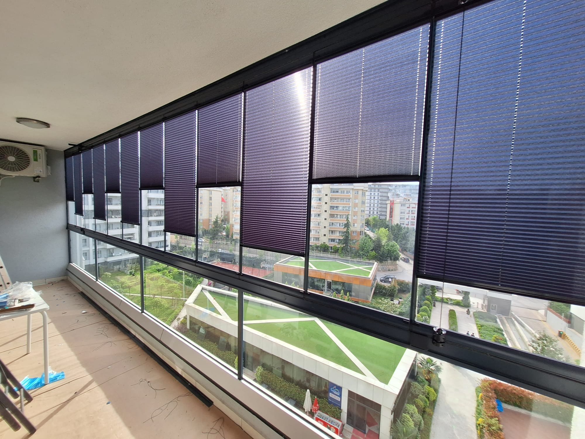 A room with wide floor-to-ceiling windows covered by dark purple blinds, overlooking a cityscape with residential buildings and a sports court below. An air conditioning unit is mounted on the upper left wall, and some furniture and construction mate