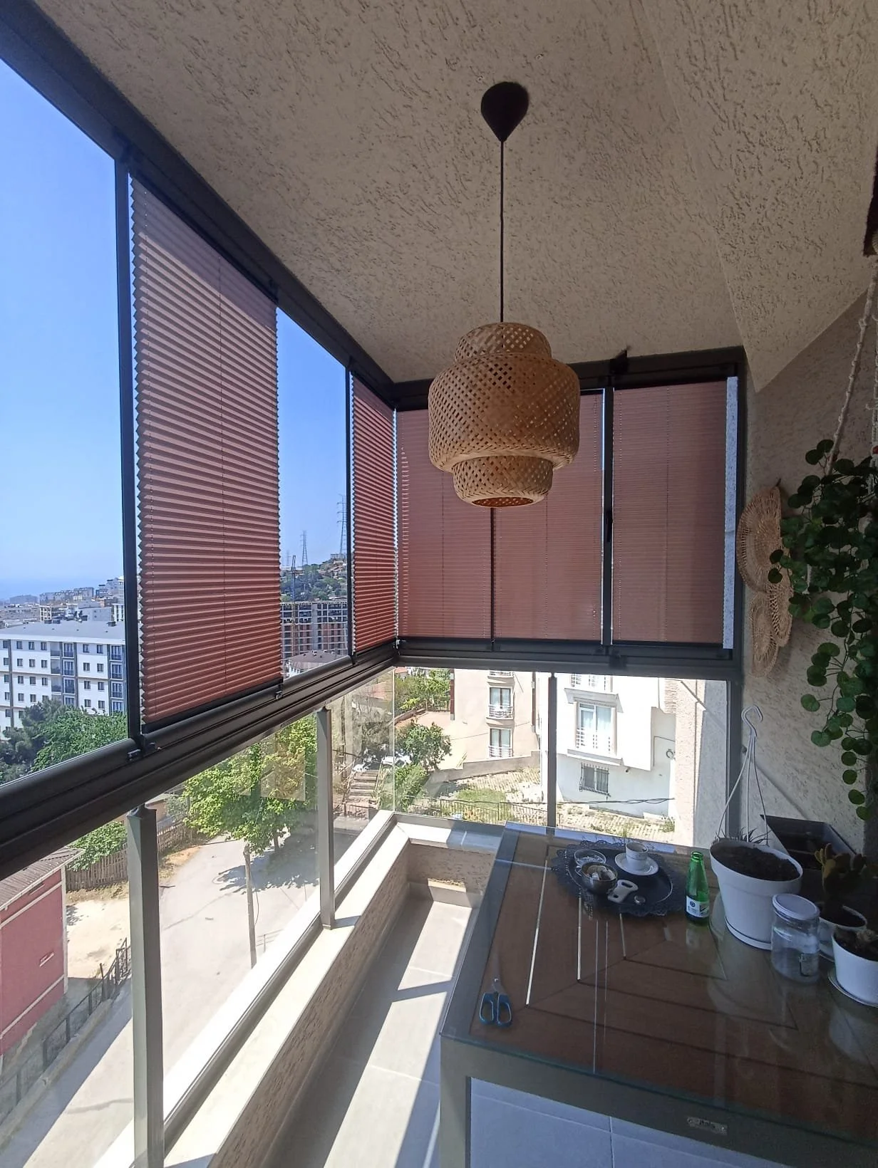 Sunlit balcony with glass windows and brown blinds, view of city buildings and sky, wicker pendant light hanging from ceiling, potted plant on corner, table with scissors, bottles, and jars.