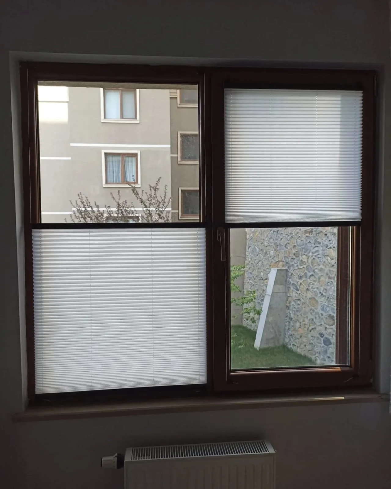 View from inside a room looking out through two windows with white blinds, showing neighboring building with gray walls, windows, and a stone wall outside.