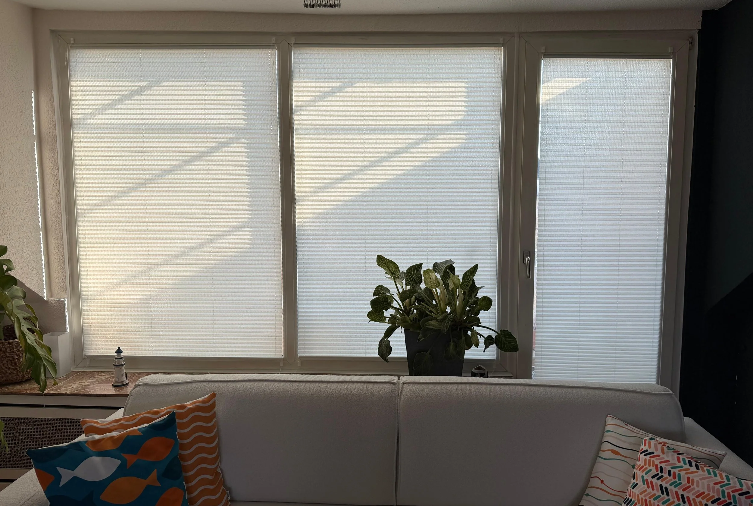 Living room view of a large window with closed horizontal blinds, a potted plant on a window sill, and colorful pillows on a white sofa.
