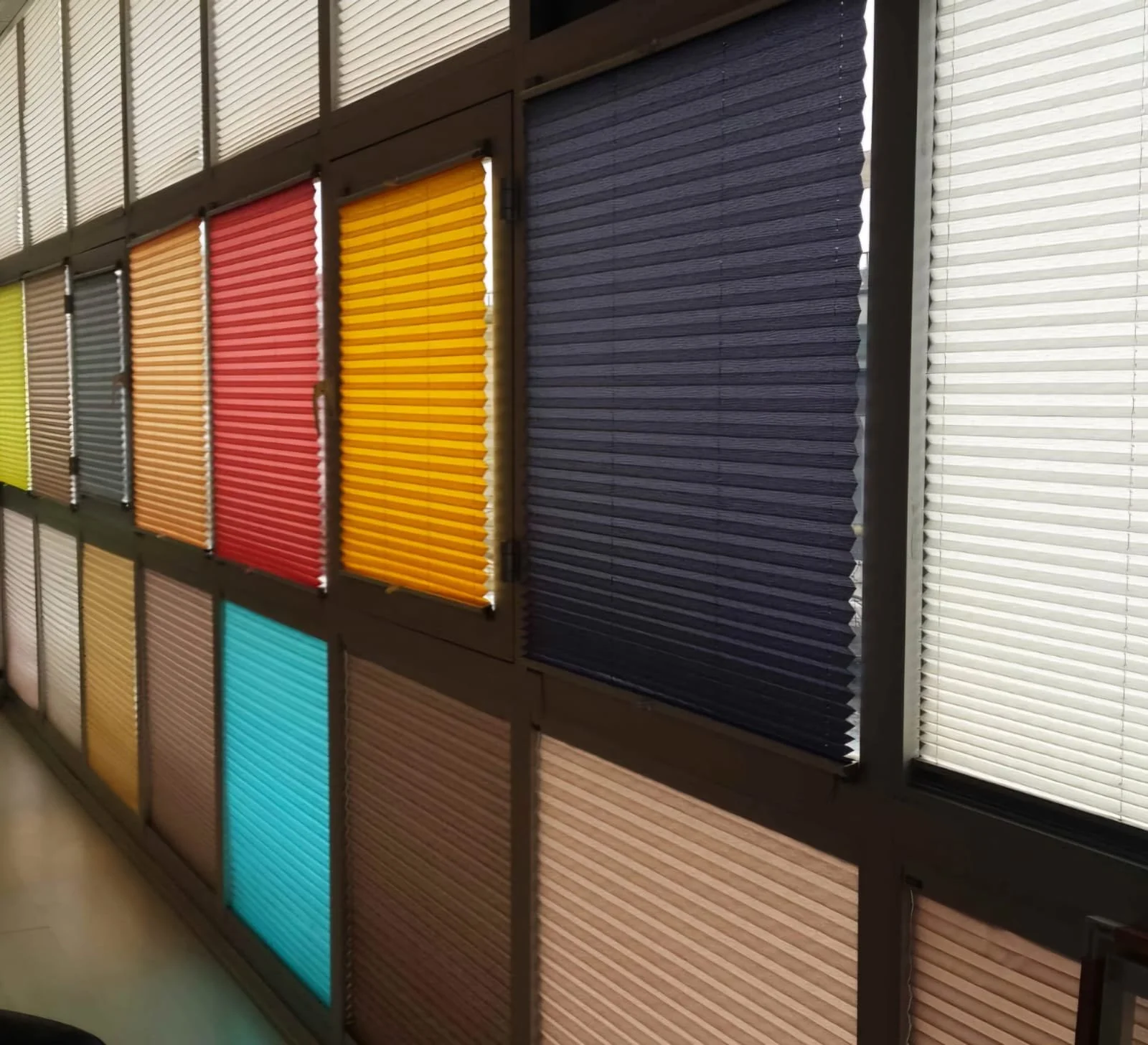 Display of colorful window blinds in various shades including blue, yellow, red, orange, and beige arranged in a grid on a wall.
