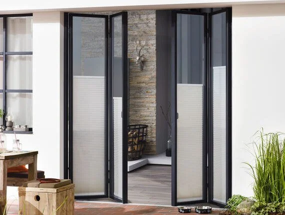 Open black-framed folding glass door leading to an outdoor patio area with a stone wall, potted plants, and outdoor furniture.