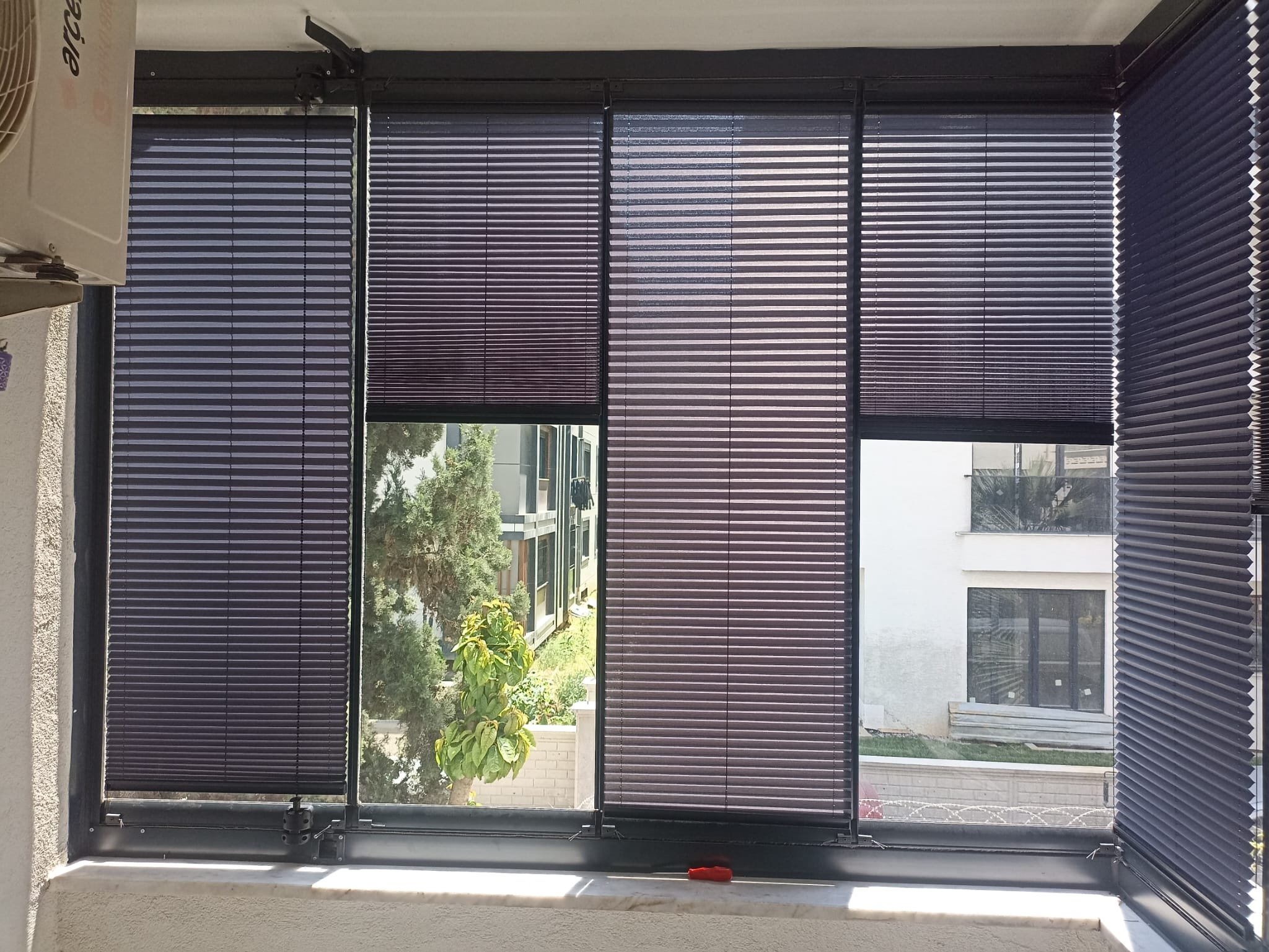 Indoor view of a window with black horizontal blinds, partially opened, overlooking a garden with plants and a building outside.