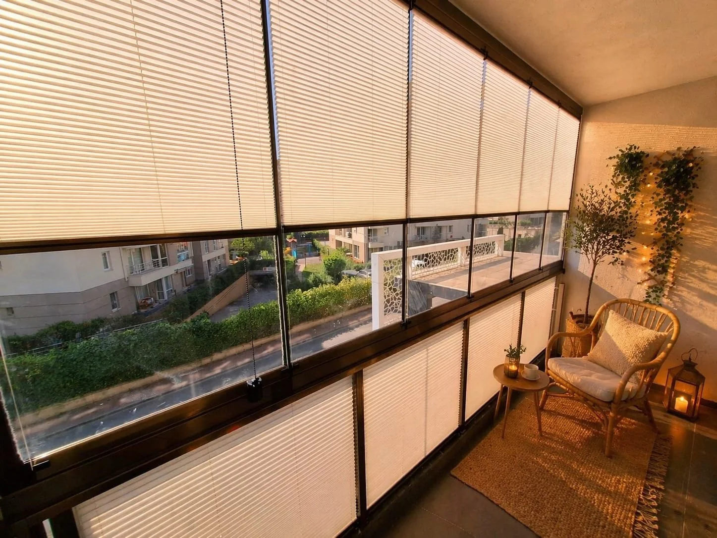 Sunlit balcony with beige blinds, a wicker armchair with a cushion, a small table with a plant and candle, potted olive tree, string lights, and lantern on the floor.