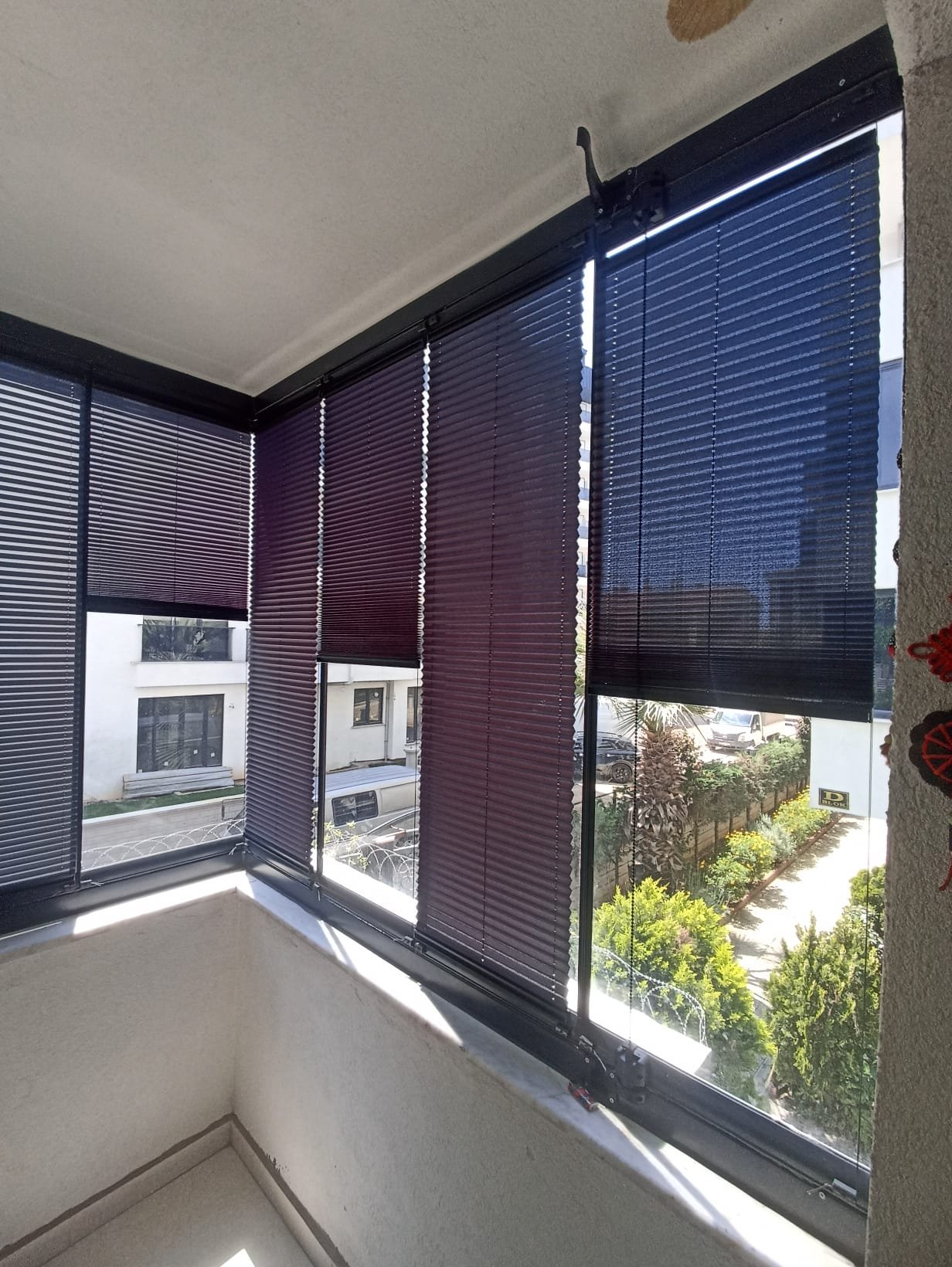Close-up of a corner of a balcony with folding window blinds in dark colors, partially open, showing a view of a street with cars, buildings, and greenery outside.