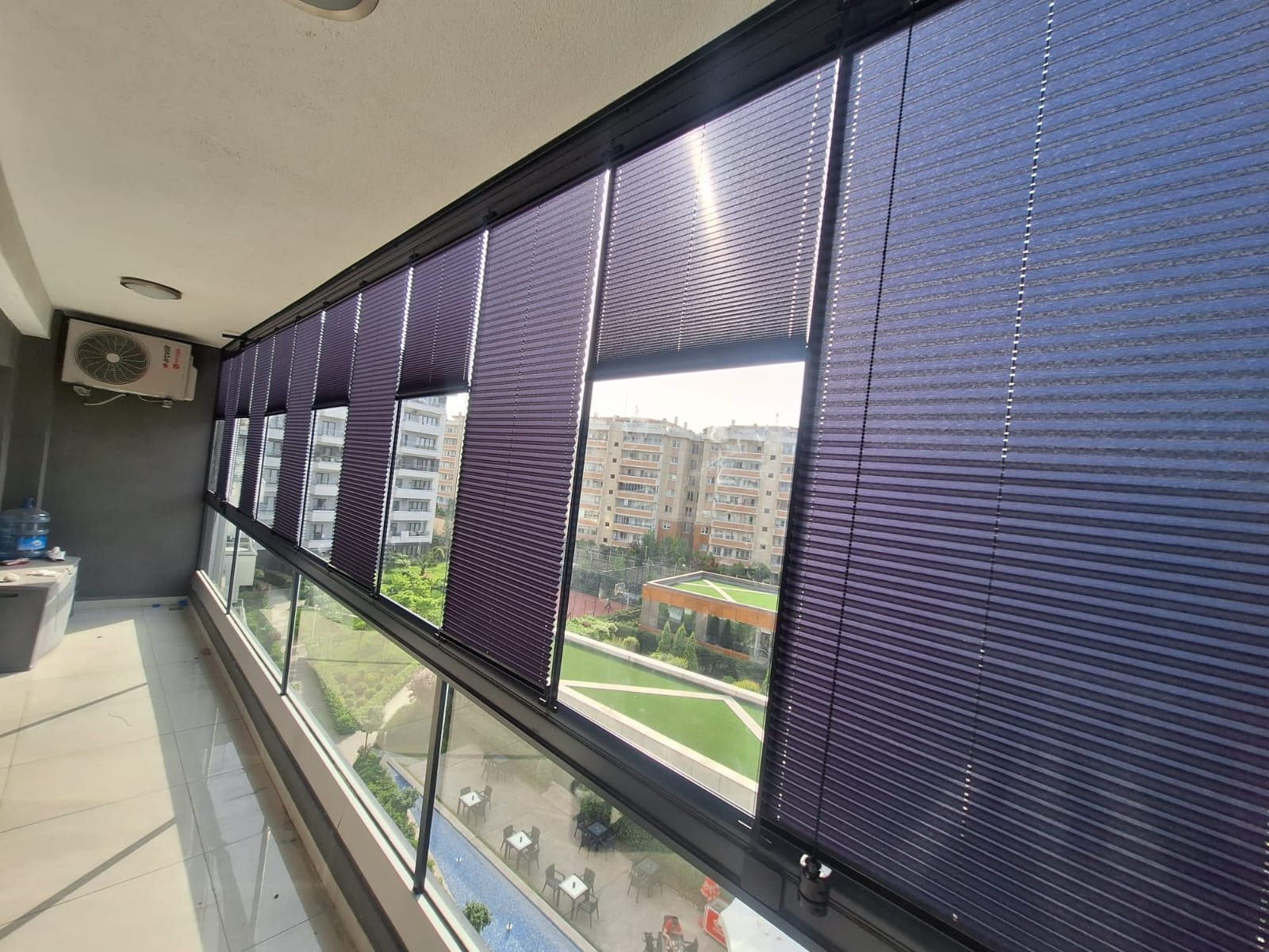 Indoor balcony with purple venetian blinds covering large windows, overlooking a residential area with modern apartment buildings and a garden.