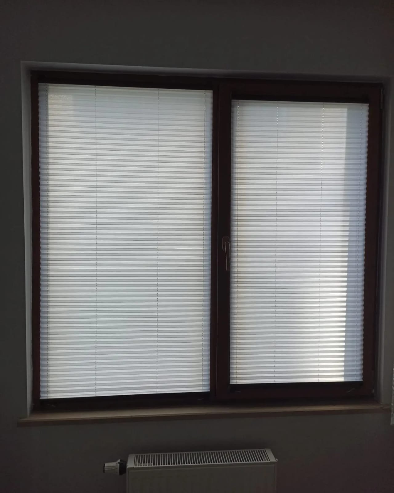 Photo of a large window with closed white blinds, a dark brown frame, and a small handle on the right side, set in a gray wall.