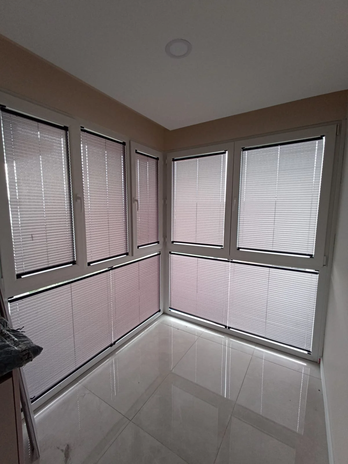 A corner room with large windows covered by closed beige window blinds, reflecting on the shiny tiled floor.