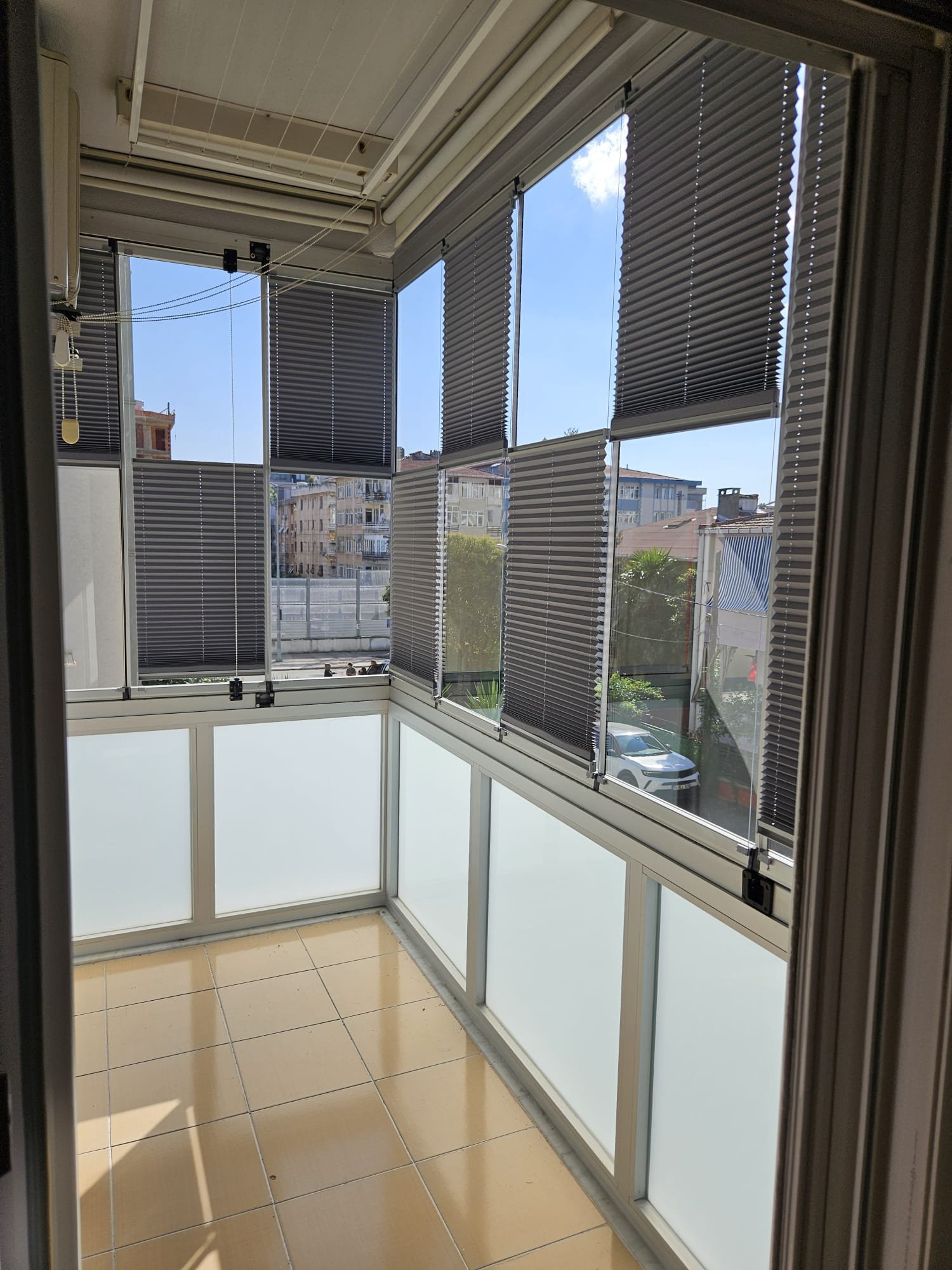 A small enclosed balcony with beige tiled flooring, large transparent windows with black horizontal blinds, and a view of neighboring apartment buildings and a street with parked cars.