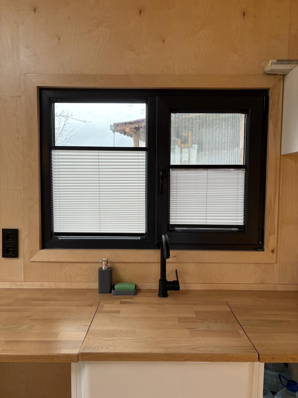 View of a kitchen window with black trim, two open window panes with blinds, black faucet, soap dispenser, and a sponge on a wooden countertop, with light wood walls.