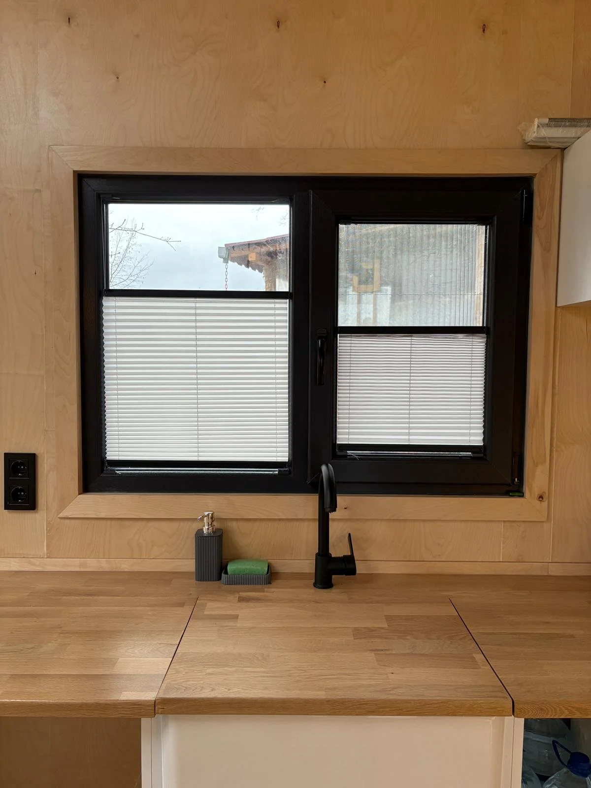 Close-up of a kitchen window with a black frame, partially covered by closed white blinds. There is a black faucet below the window, two electrical outlets on the left wall, and a soap dispenser along with a green scrub sponge on the wooden counterto