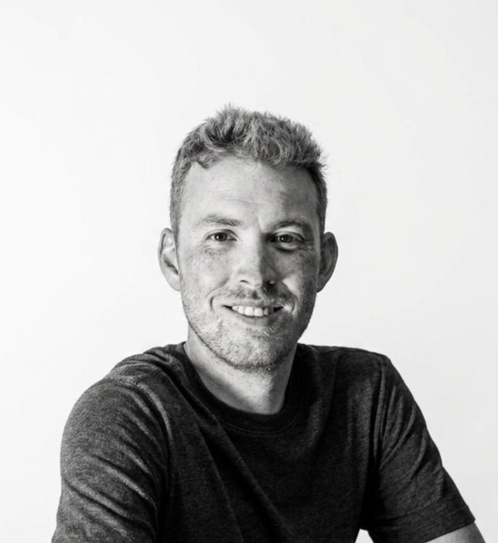 Black-and-white portrait of a young man with short, light-colored hair, smiling and wearing a dark t-shirt, against a plain light background.