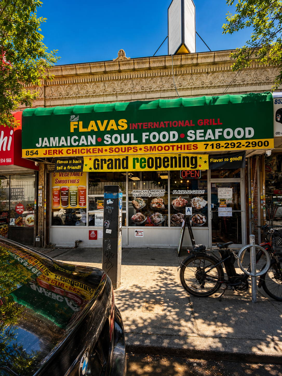 Front of a Jamaican restaurant with a green awning announcing a grand reopening. The signs advertise Jamaican soul food, seafood, jerk chicken, soups, and smoothies. There is a small display with images of food and a bicycle parked outside.