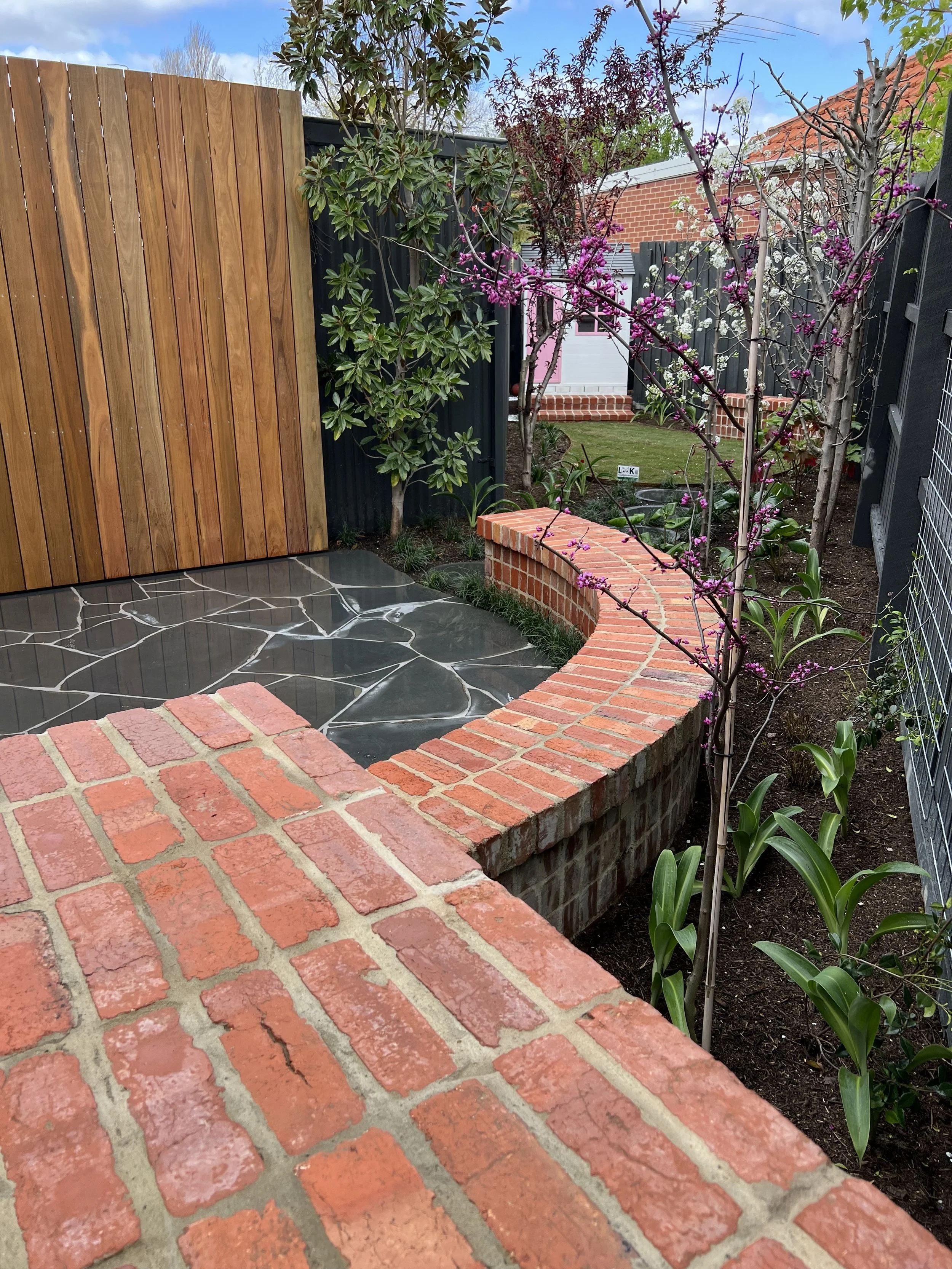 A decorative garden with a brick and stone patio, a curved brick border, a wooden fence, various trees and plants, and a white house in the background.