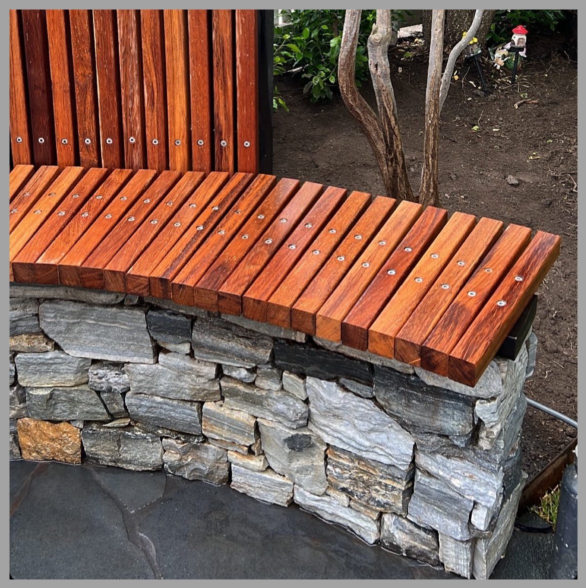 A curved stone bench with a wooden slat backrest and seat, located outdoors near a dirt garden and small trees.