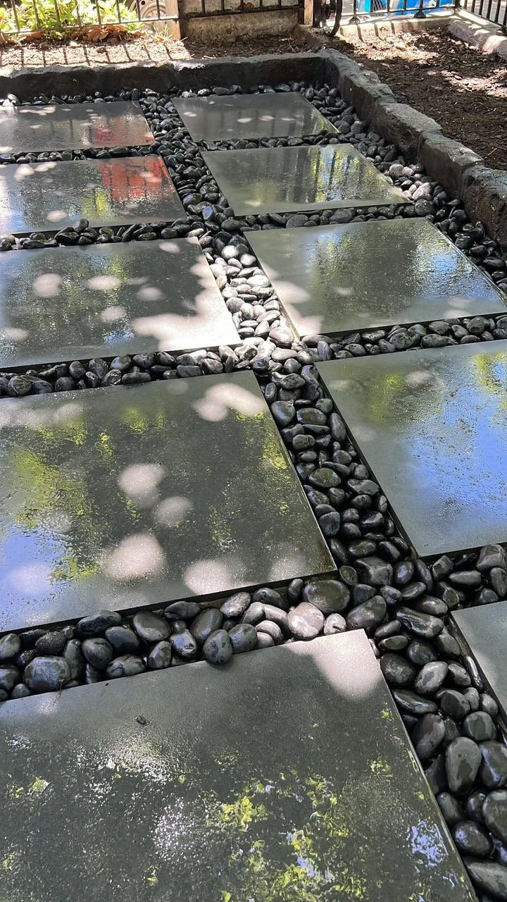 Square concrete stepping stones with reflective surfaces, separated by black smooth pebbles, under a partly shaded outdoor setting.