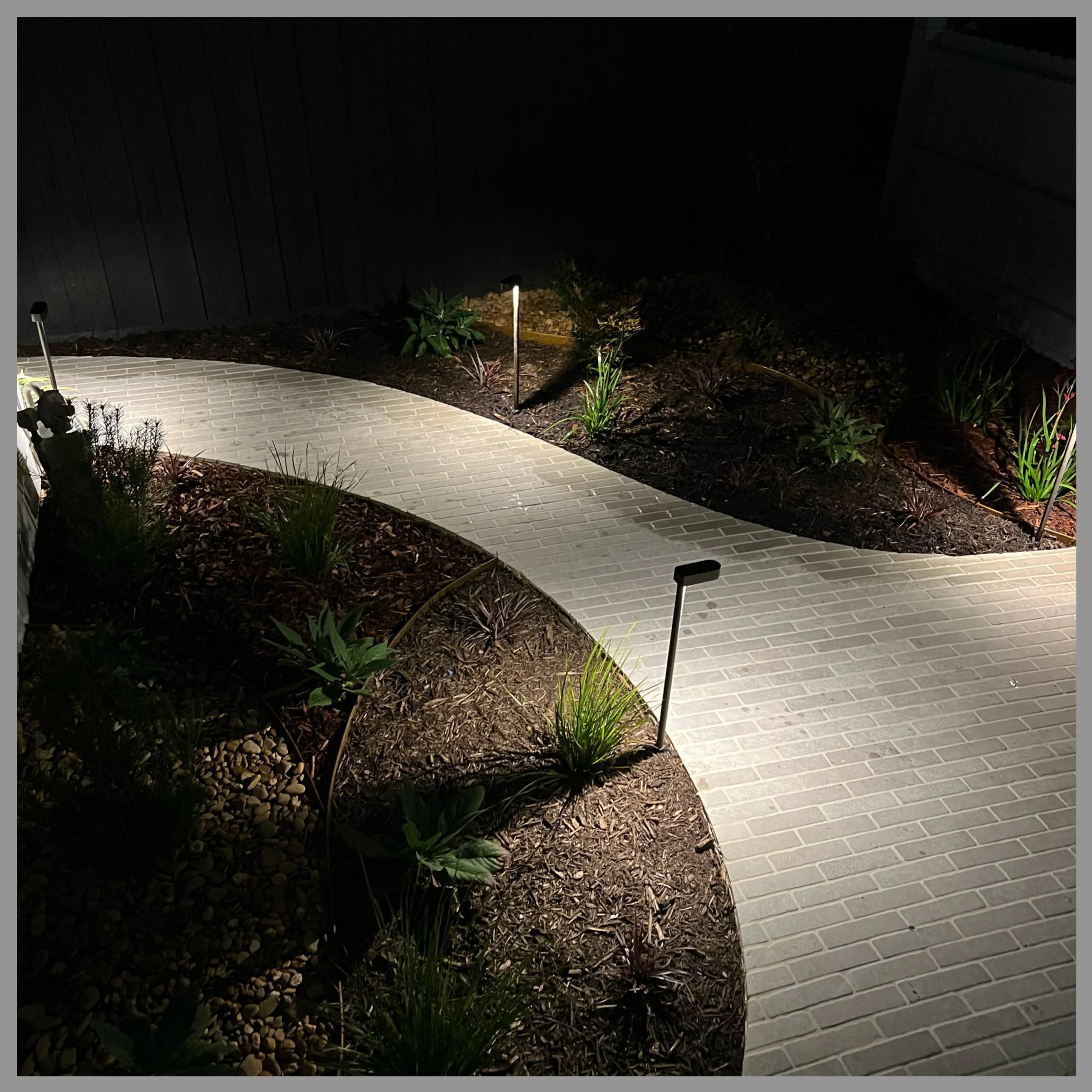 A backyard garden at night with a brick pathway illuminated by outdoor lighting. There are plants and shrubs along the edges of the pathway, with a dark fence in the background.