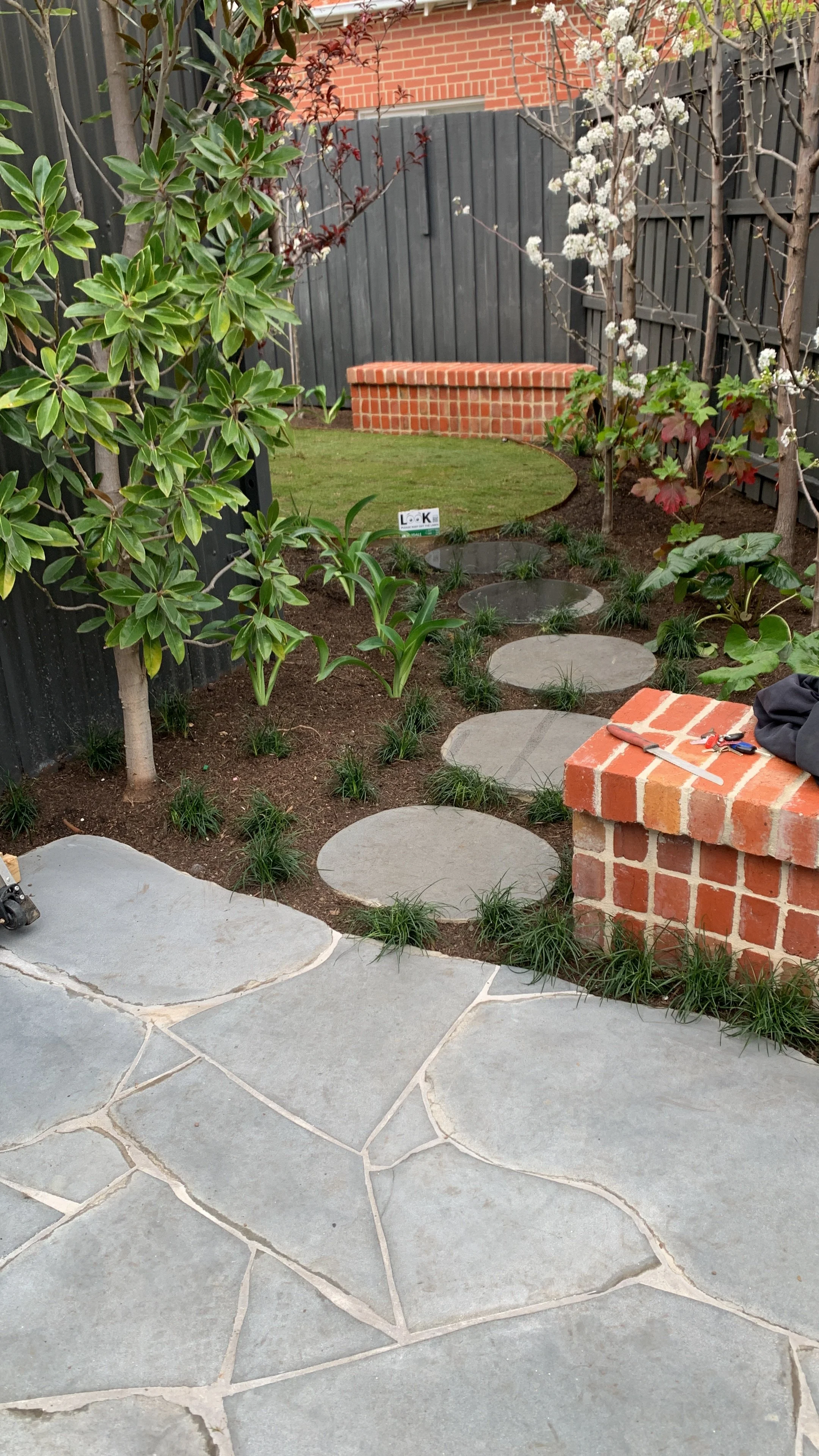 A backyard garden with stone and concrete stepping stones creating a pathway. There are plants and trees along the sides, a brick wall in the background, and a small patch of grass with a decorative brick border. Gardening tools and keys are placed on a brick ledge.