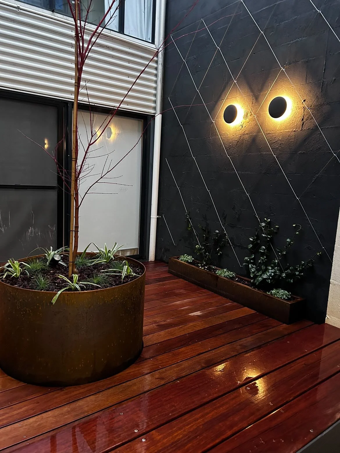An outdoor patio area with wooden flooring, a large potted plant with a small tree, a rectangular planter with plants, a black wall with decorative lines, and two circular wall lights emitting warm yellow light.