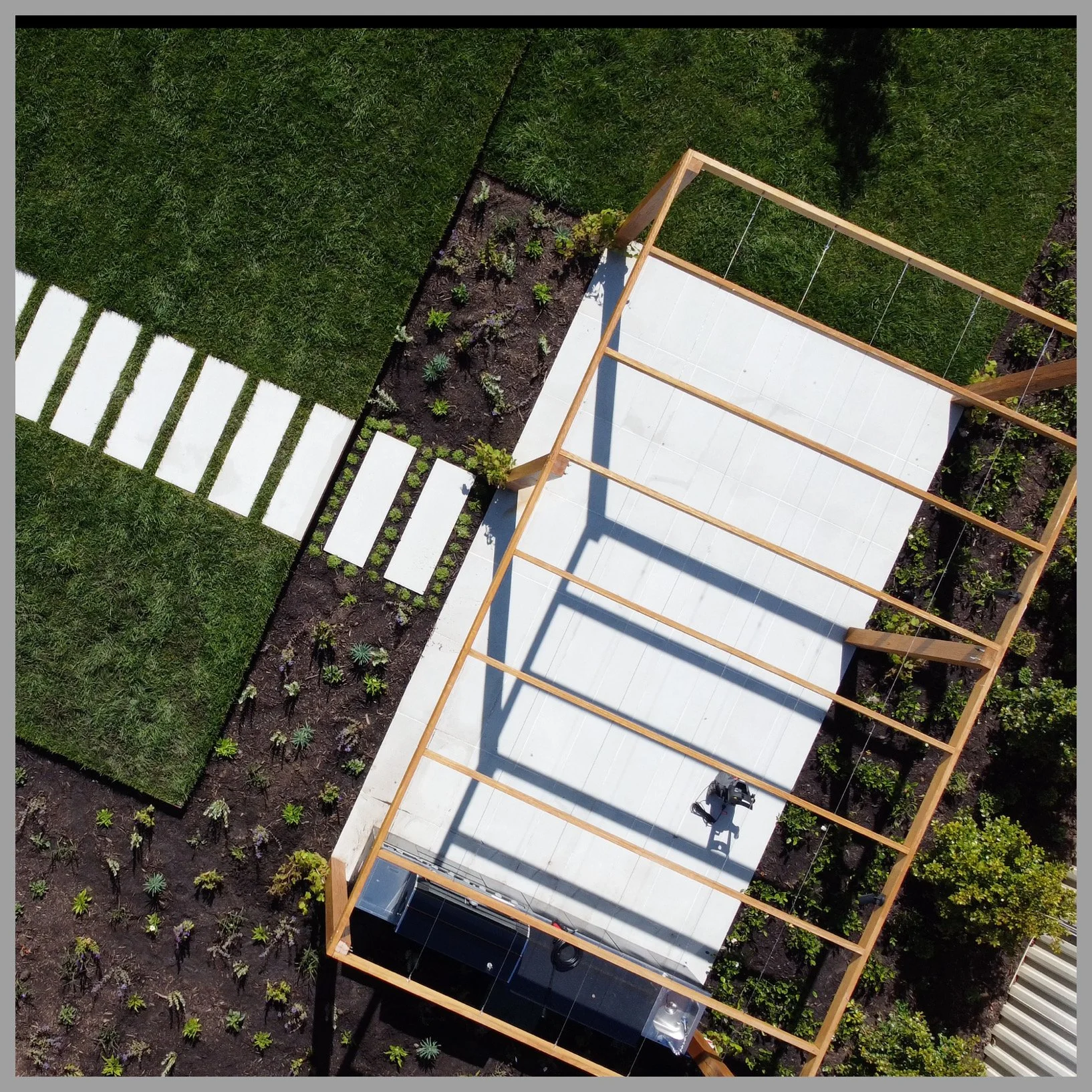 Aerial view of a garden with a partially constructed wooden frame structure, a concrete patio, a small garden bed with plants, and a walkway with white stones surrounded by green grass.