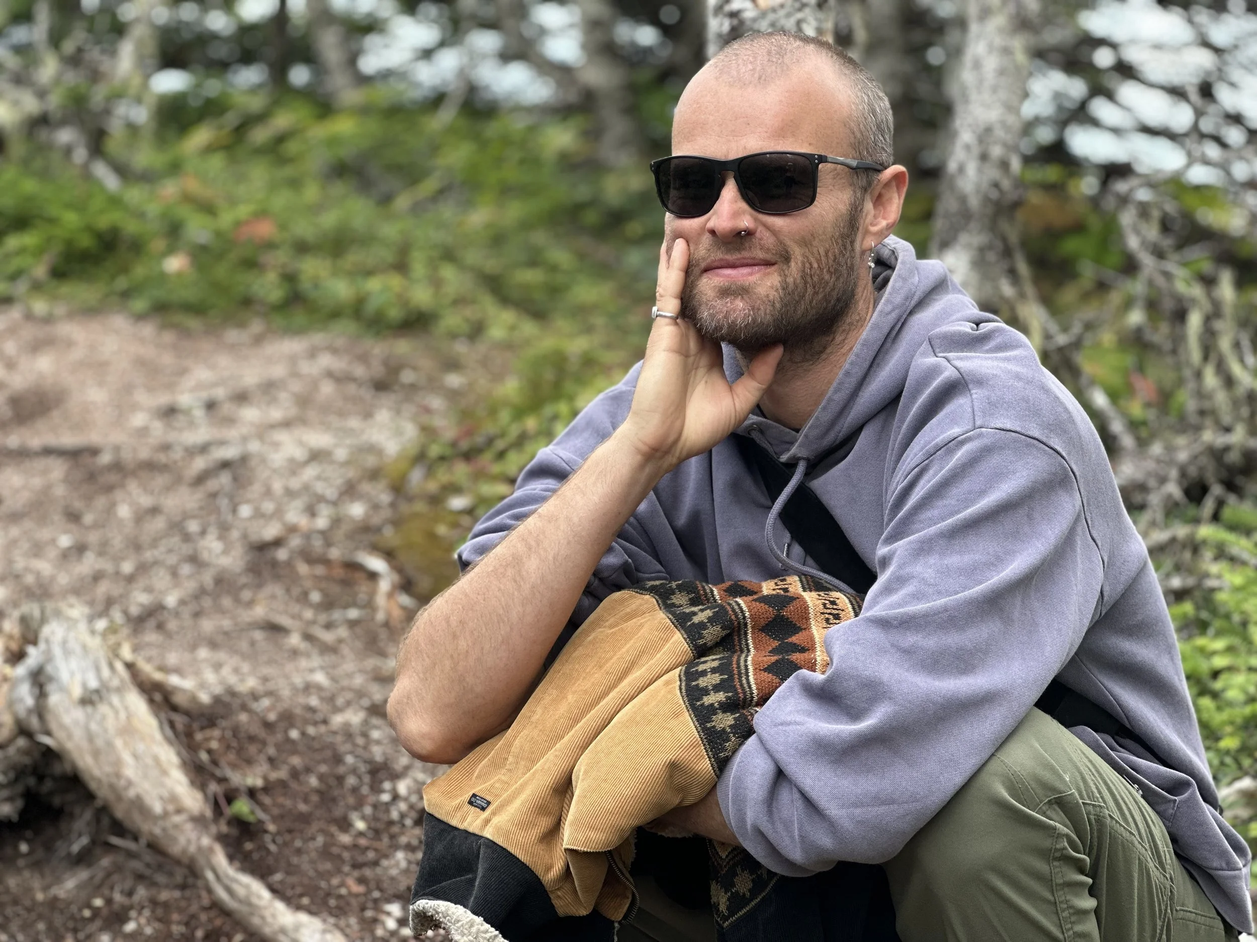 A man wearing sunglasses, a gray hoodie, and tan pants outdoors in a wooded area, resting his head on his hand near a fallen tree.
