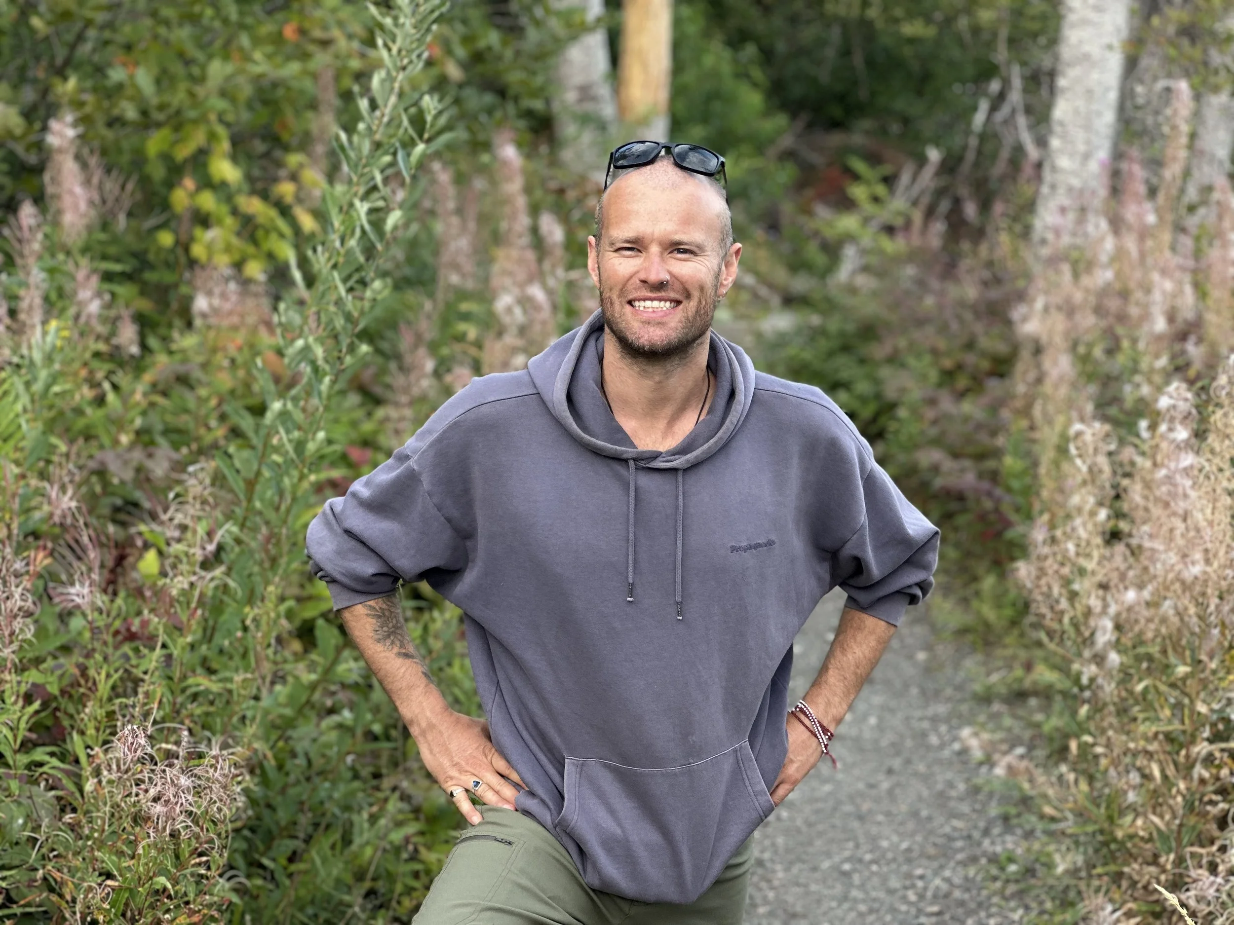 A smiling man with sunglasses on his head, wearing a gray sweatshirt and green pants, standing outdoors on a nature trail surrounded by green foliage and tall trees.