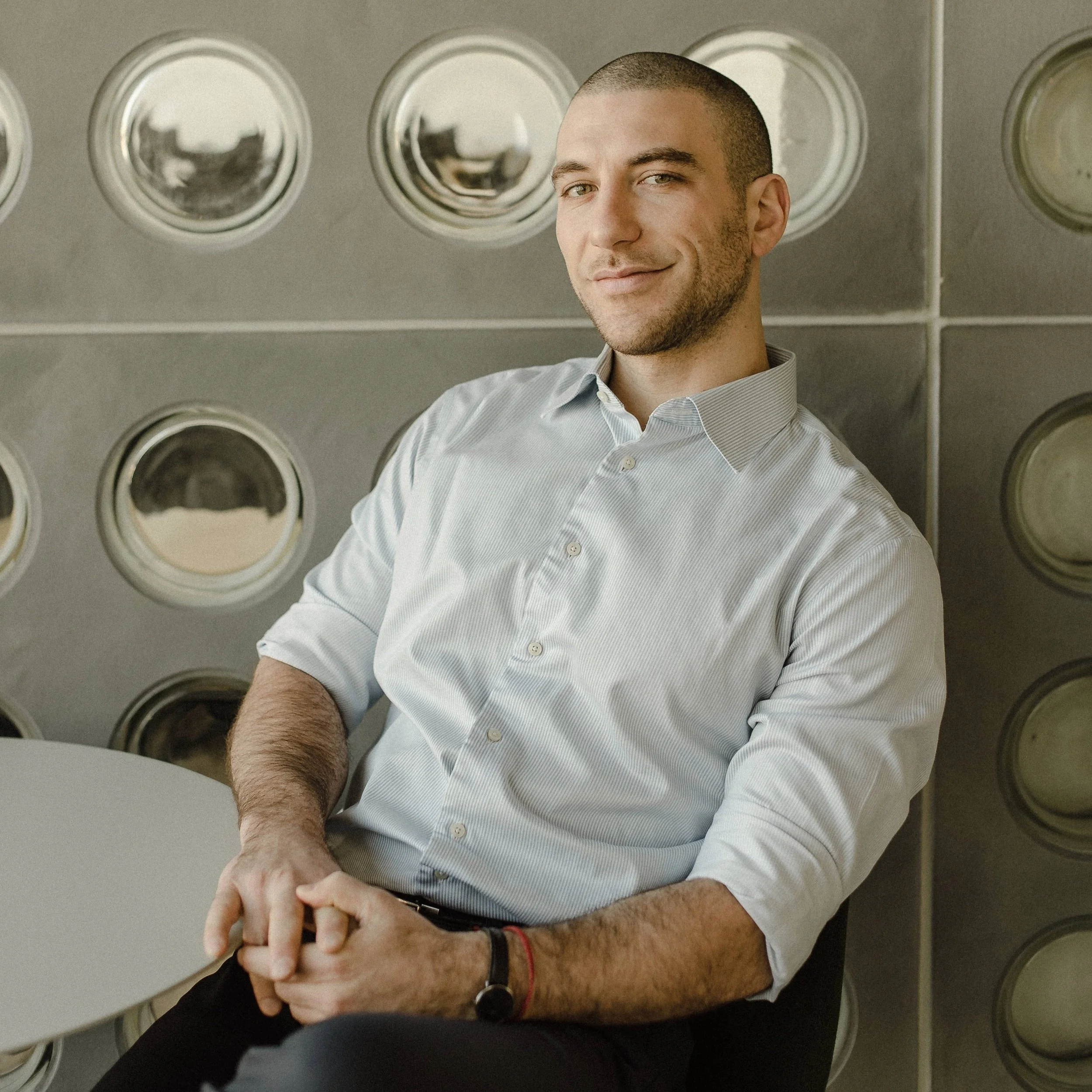 A young man with short hair, wearing a light blue button-up shirt, smiling slightly, standing against a wall with reflective circular panels.