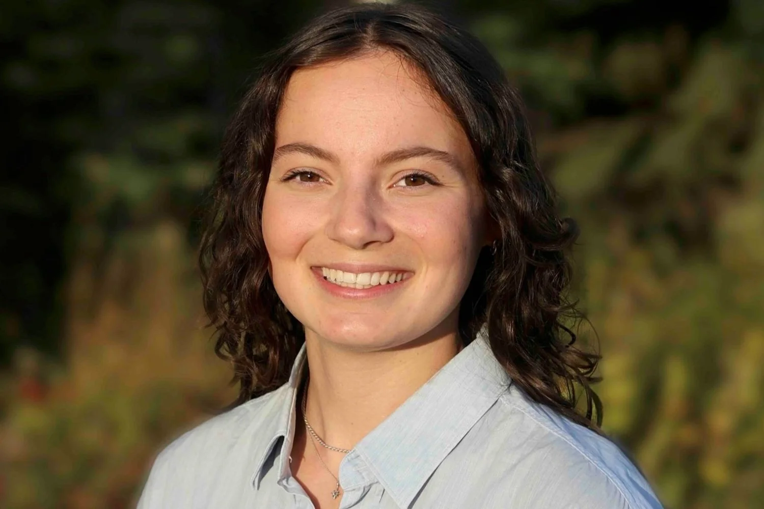 Close-up of a woman with shoulder-length curly brown hair, smiling, wearing a light-colored button-up shirt, outdoors with blurred green foliage in the background.