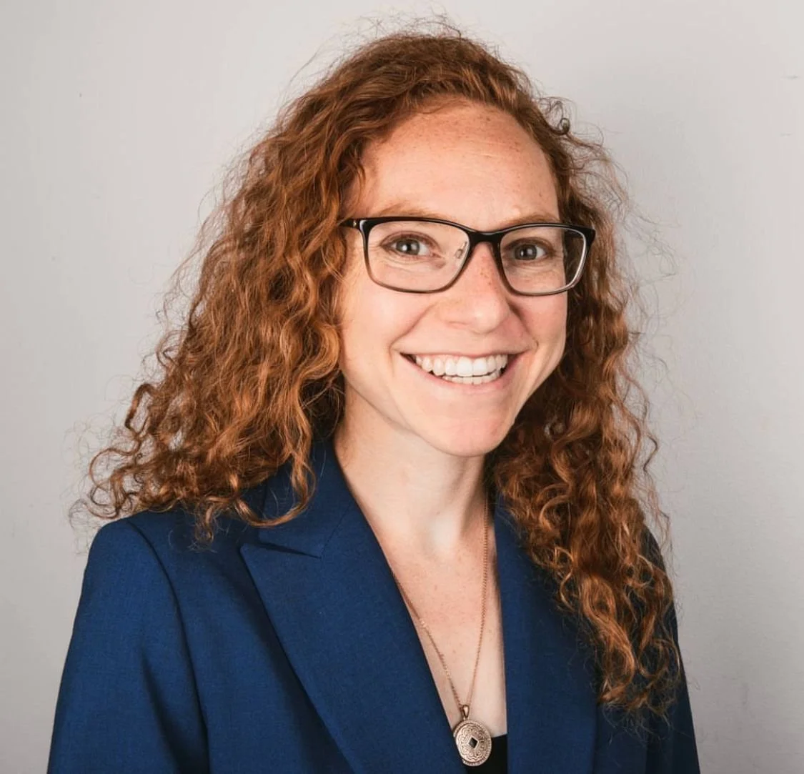 Smiling female lawyer with curly red hair, glasses, and a blue blazer, posing against a neutral background.