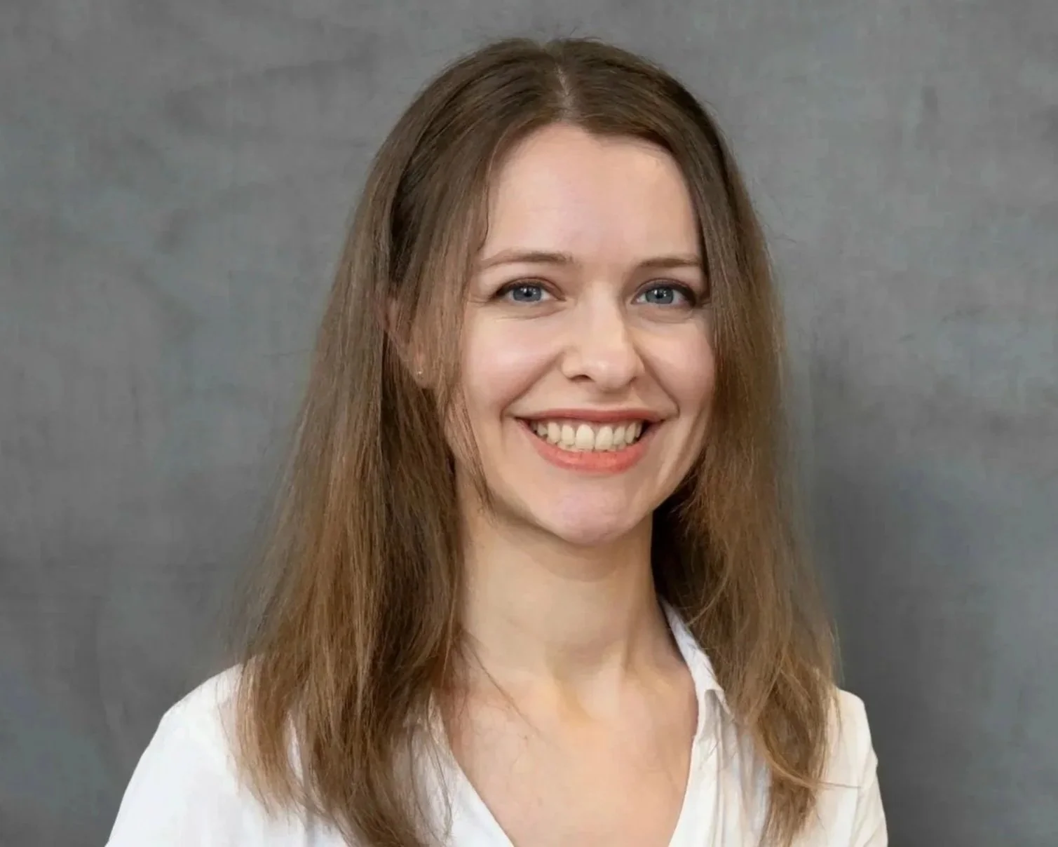 A smiling woman with long light brown hair, blue eyes, and fair skin, wearing a light-colored top, standing against a gray background.
