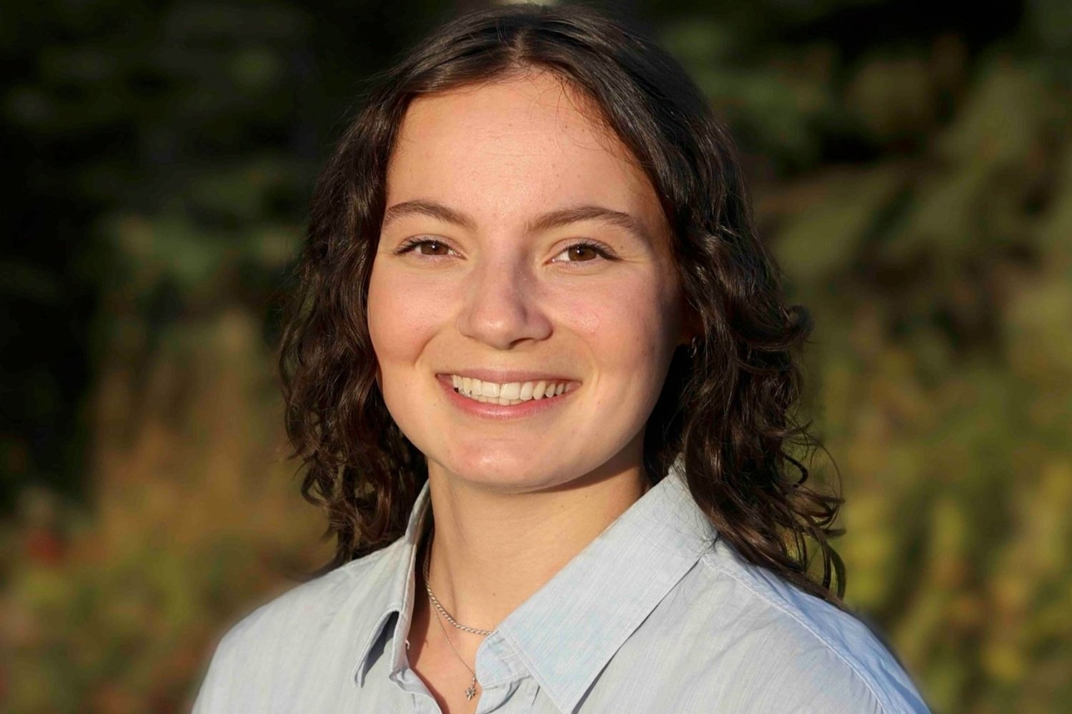 A young woman with short, curly brown hair smiling outdoors with greenery in the background.