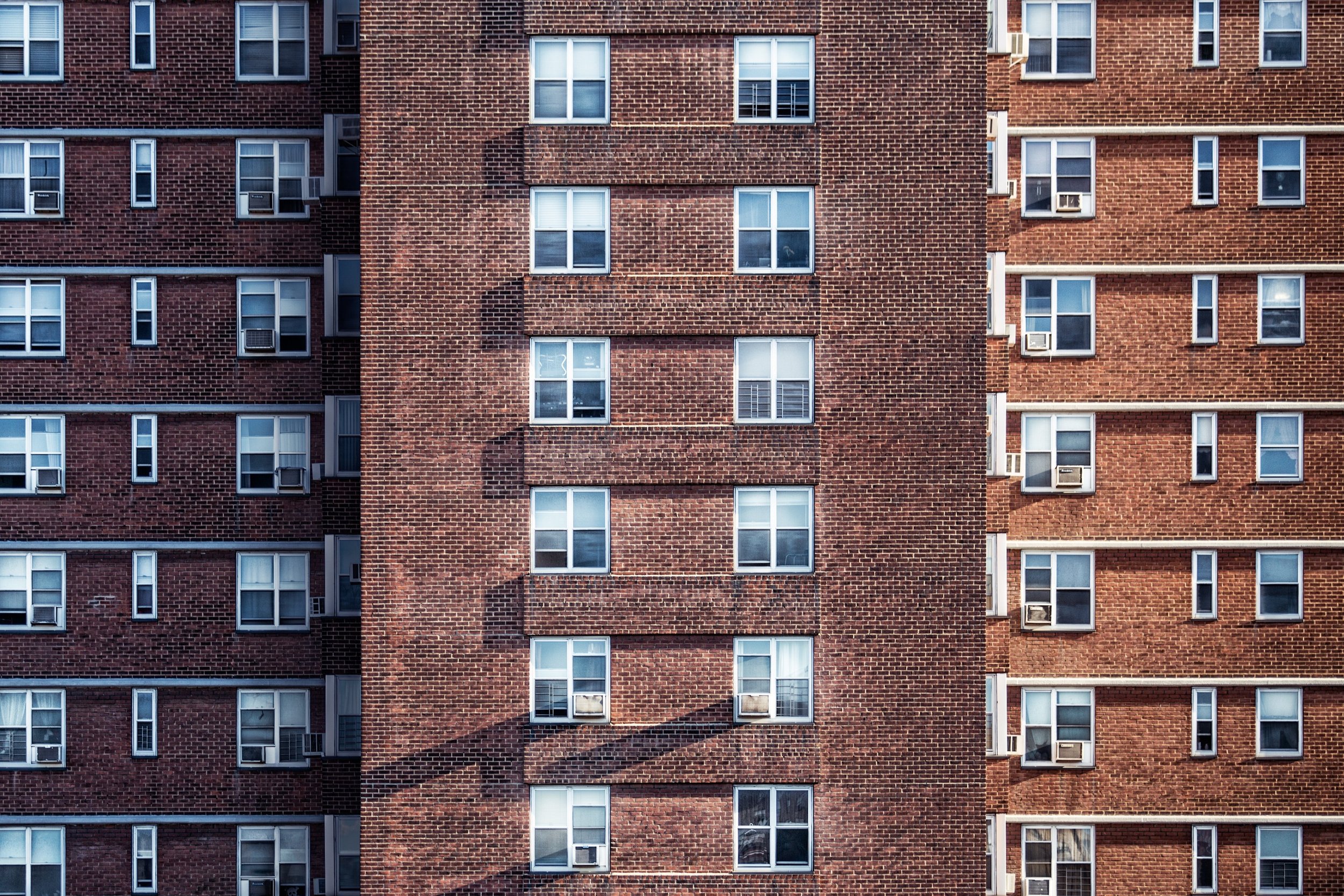 A brick apartment building with multiple windows, some of which have air conditioning units and window blinds. A shadow of a person is cast on the building's facade.
