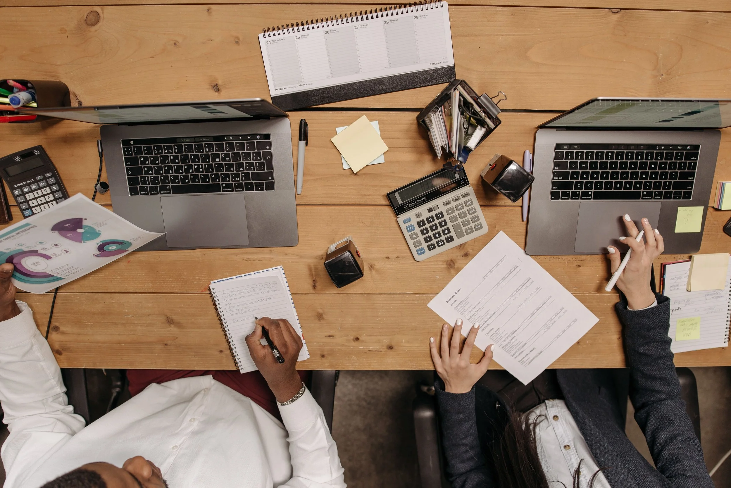 Wooden desk with laptops, papers, other office supplies, and two people working.