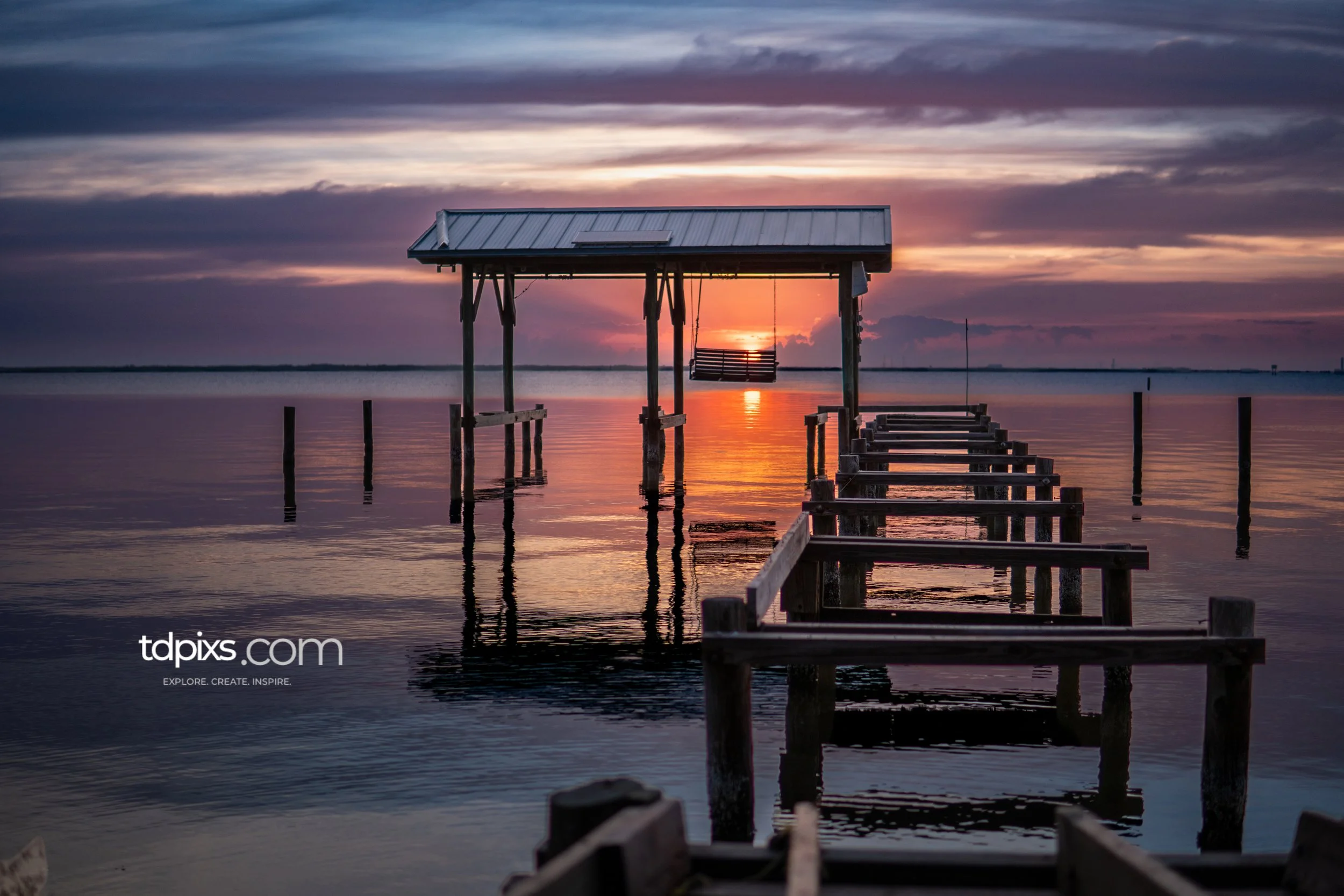 A wooden dock extending into calm water with a covered seating area, during a colorful sunset.