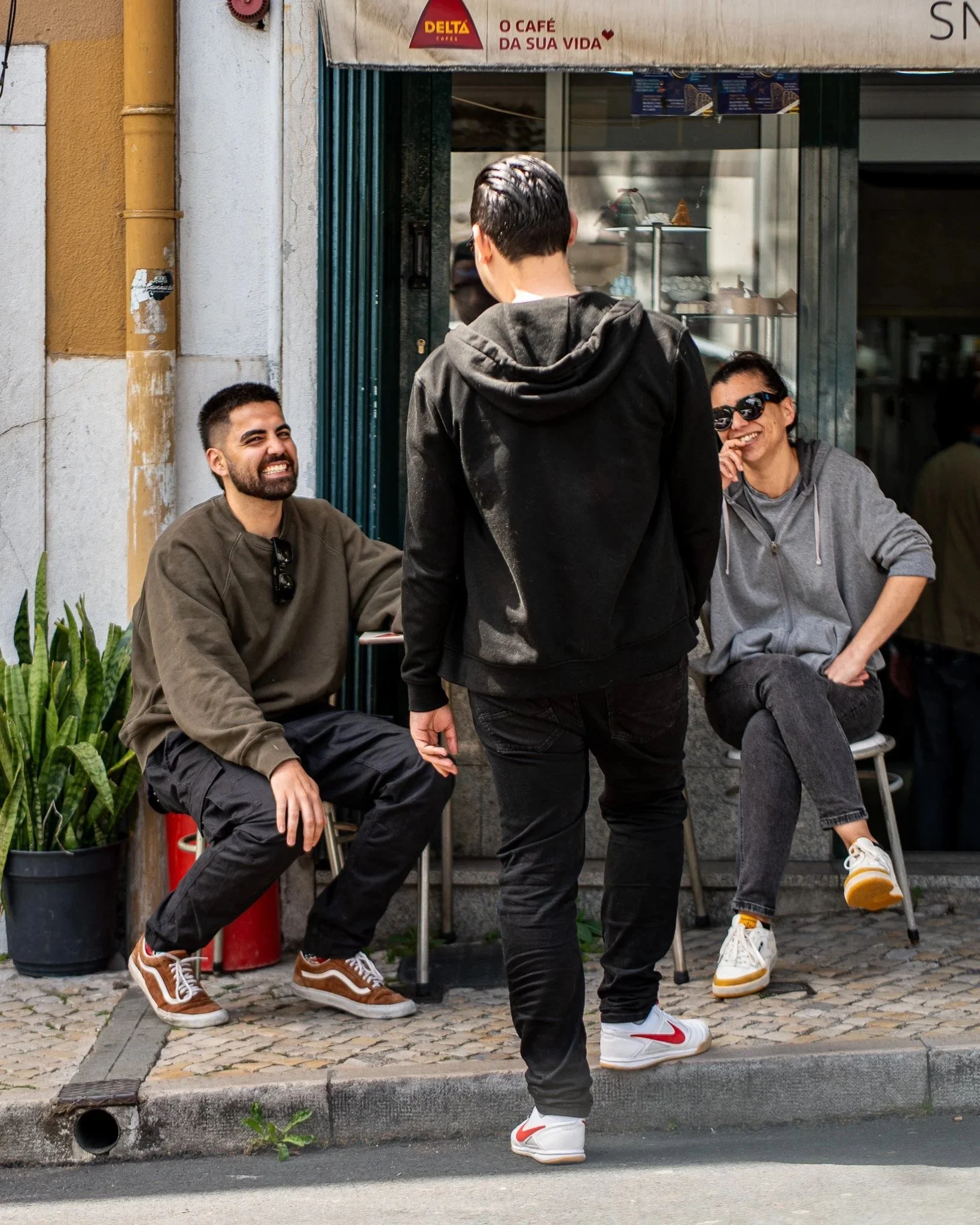 Three young men, one wearing a black hoodie, and two sitting outside a small cafe, engaging in conversation on a cobblestone sidewalk with a potted plant nearby. The cafe has signage in a foreign language and a display window with desserts inside.