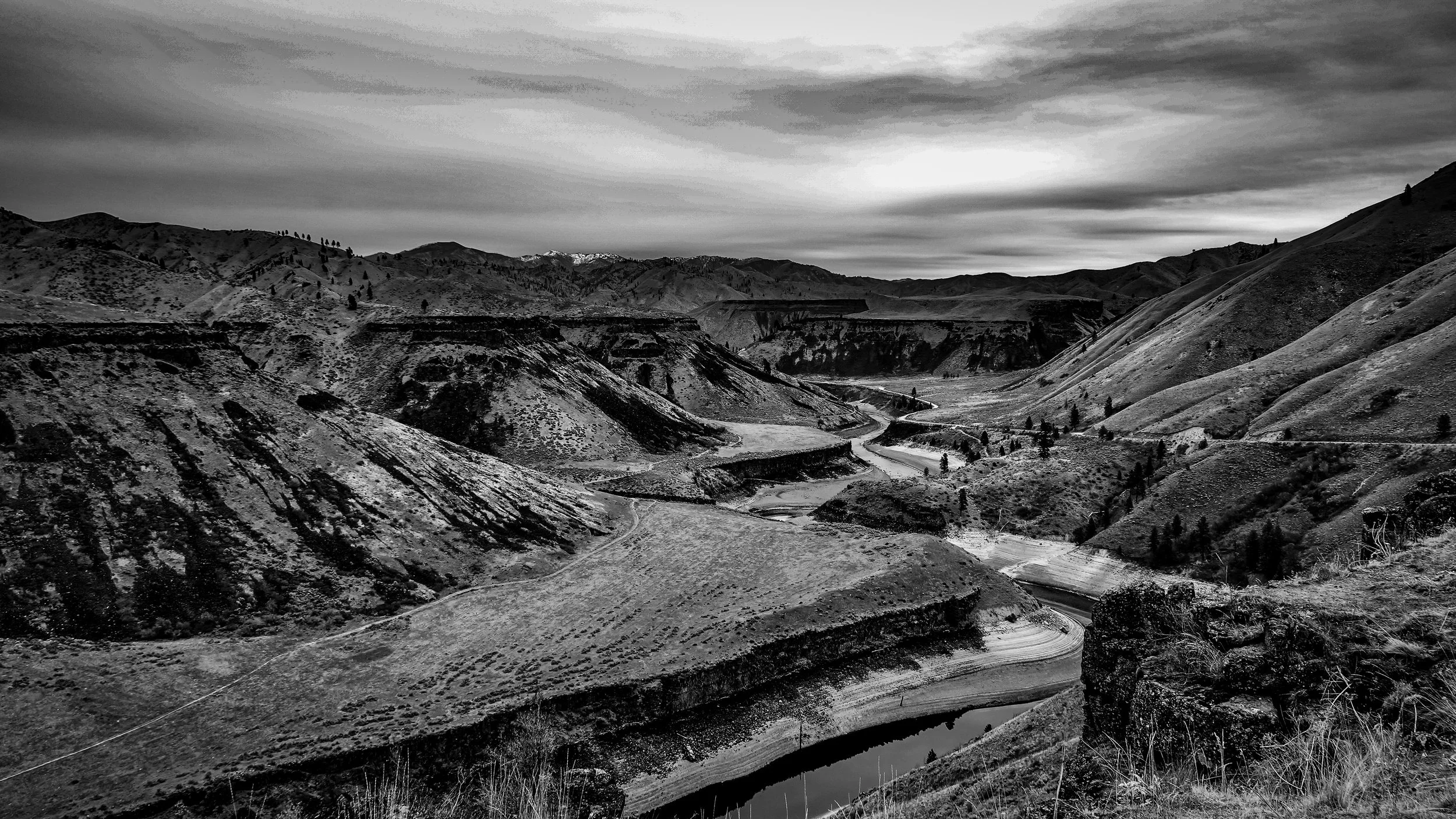 Black and white landscape photo of rolling hills, a winding river, and cloudy sky.