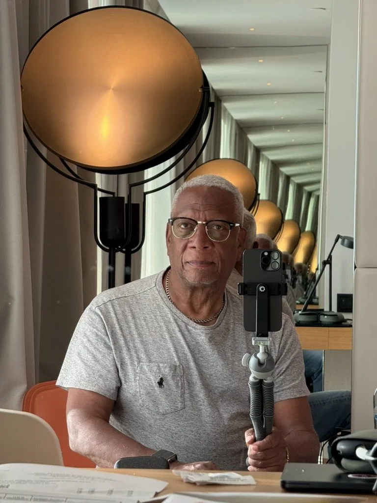 Older man with gray hair and glasses taking a selfie with a smartphone mounted on a selfie stick in a mirror-lined room with warm lighting and modern decor.