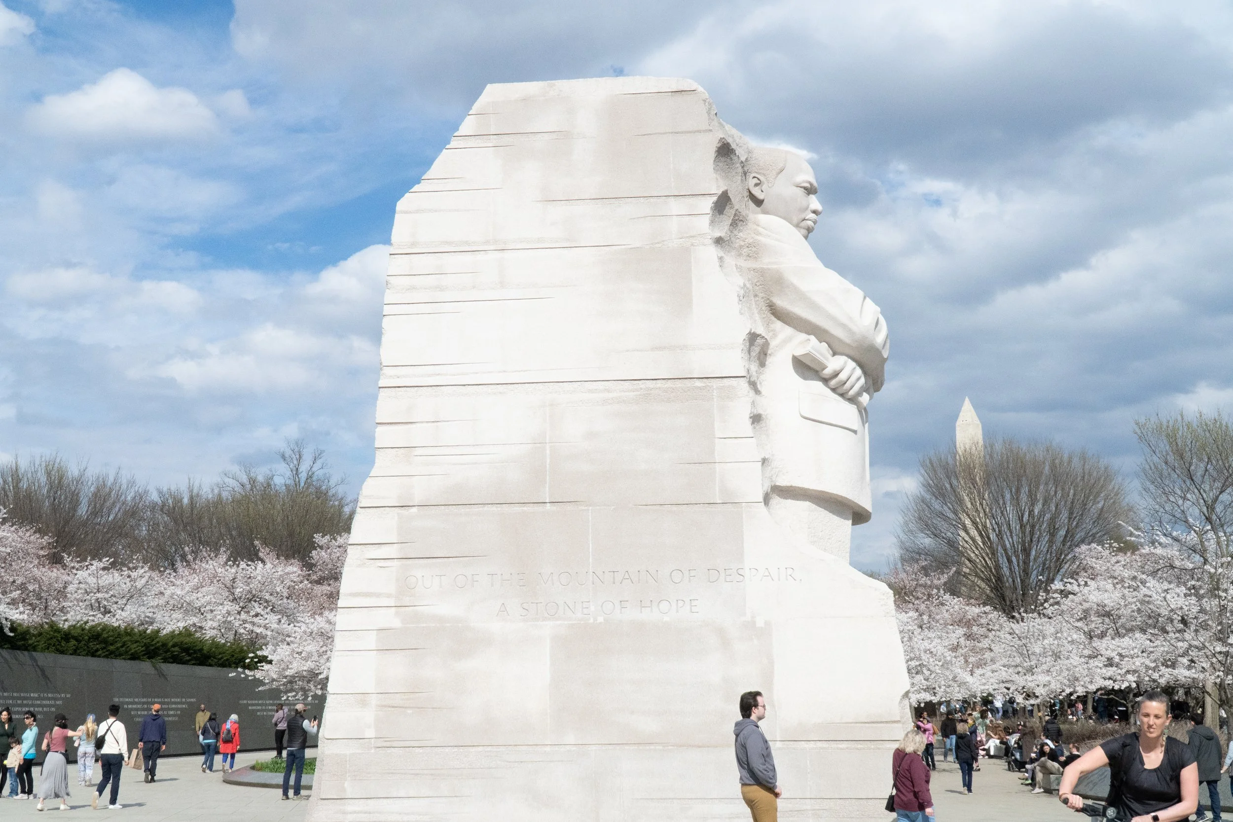 The Mount Rushmore monument with four carved faces, surrounded by cherry blossom trees and visitors at the site under a partly cloudy sky.