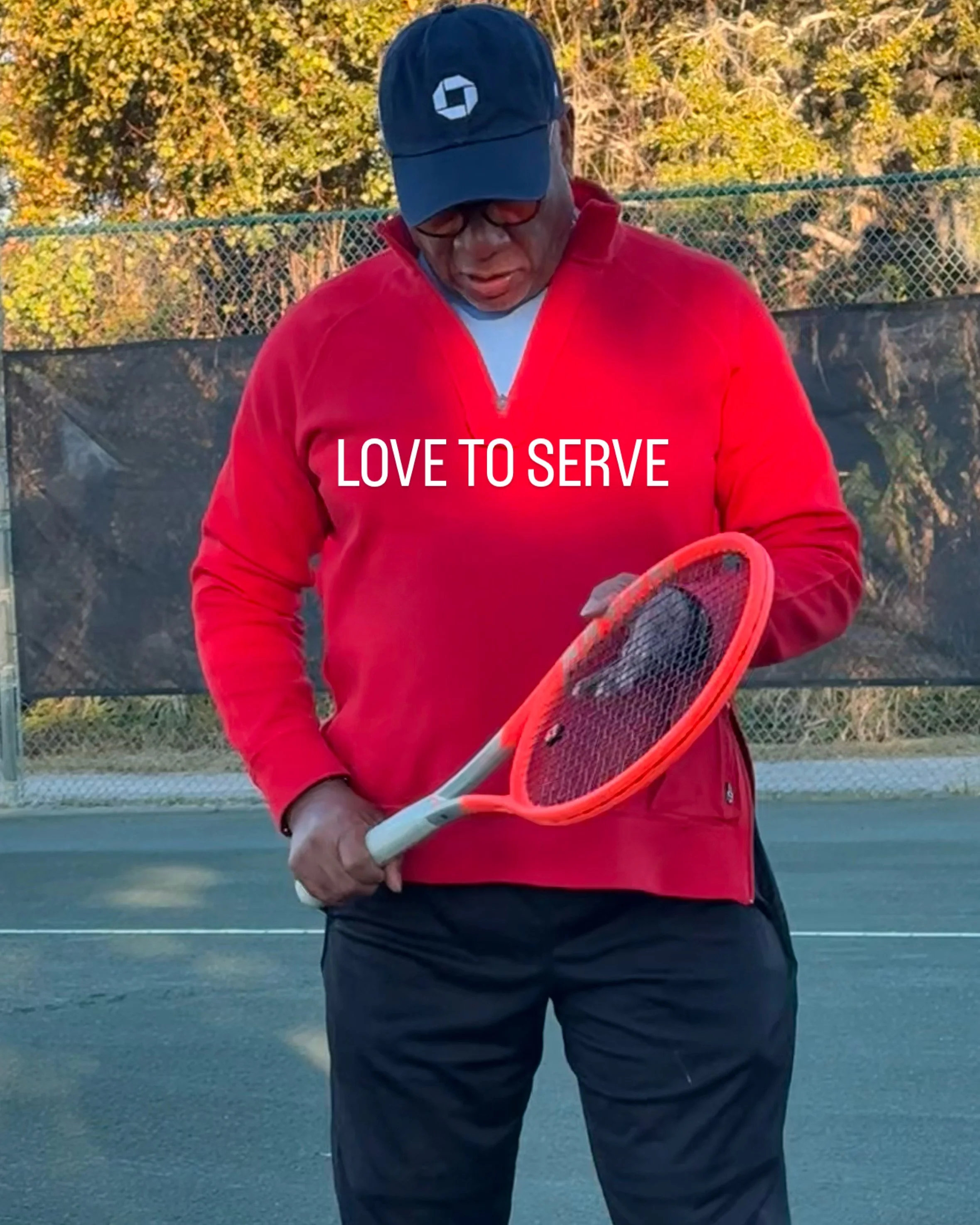 A man wearing a red jacket and black pants holding an orange tennis racket on an outdoor tennis court with trees in the background.