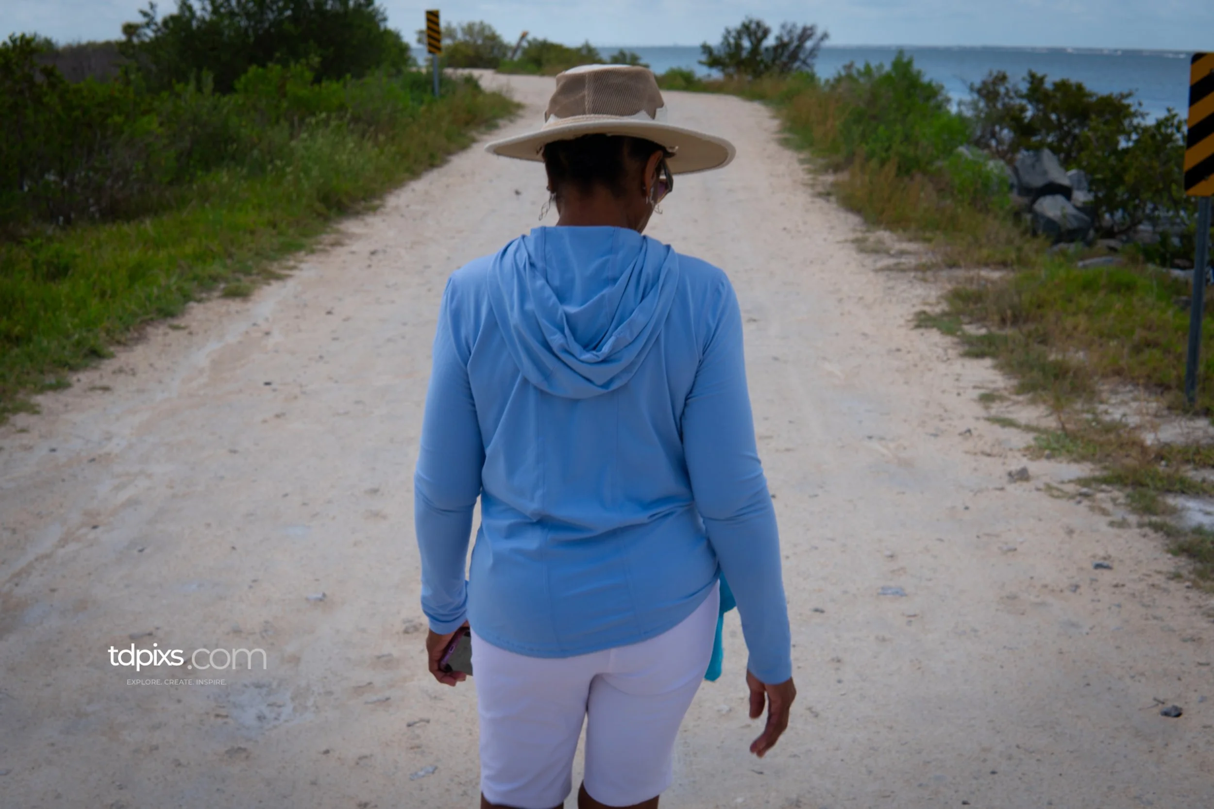 A woman walking on a dirt path near a body of water, wearing a wide-brimmed hat, sunglasses, a light blue jacket, and white pants.