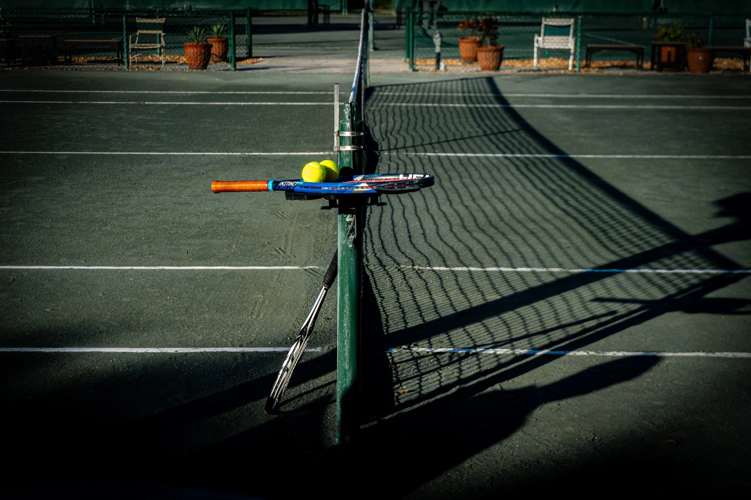 A tennis court with a tennis racket and two tennis balls resting against a green pole near the net.